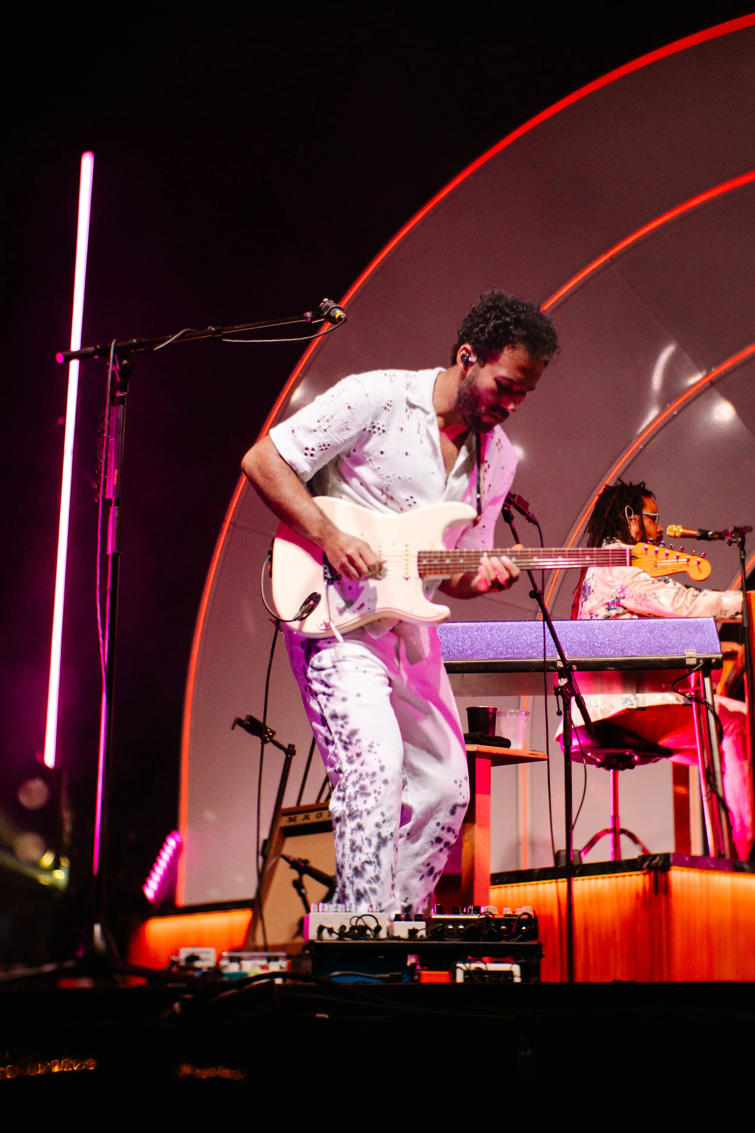 Musicians performing on stage, one playing an electric guitar and the other at keyboard, with colorful lighting and a modern stage design.