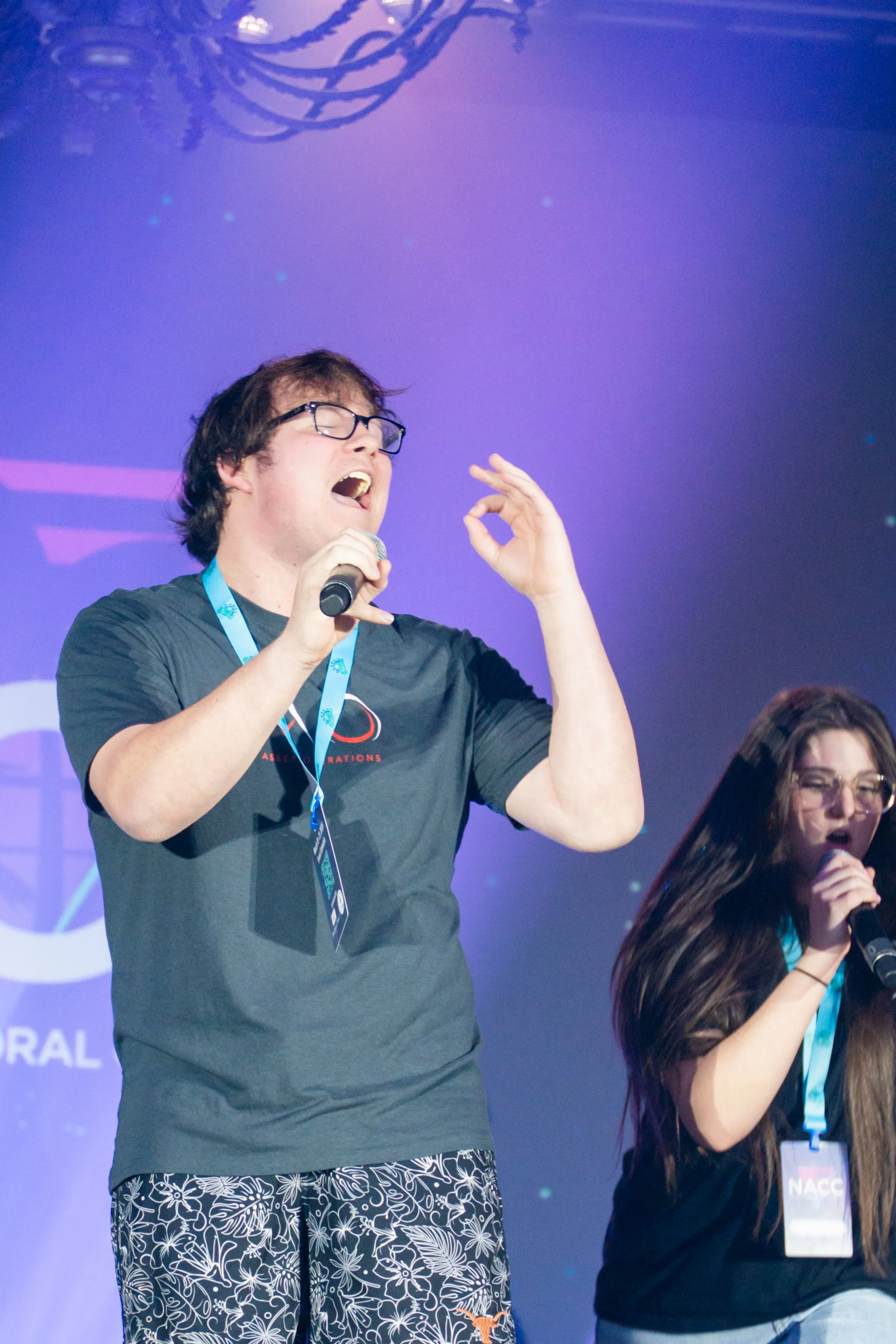 A man with dark glasses singing into a microphone on stage, wearing a black t-shirt and patterned shorts. There is a woman next to him also singing, wearing a lanyard and black top. The background is purple with abstract lights and shapes.