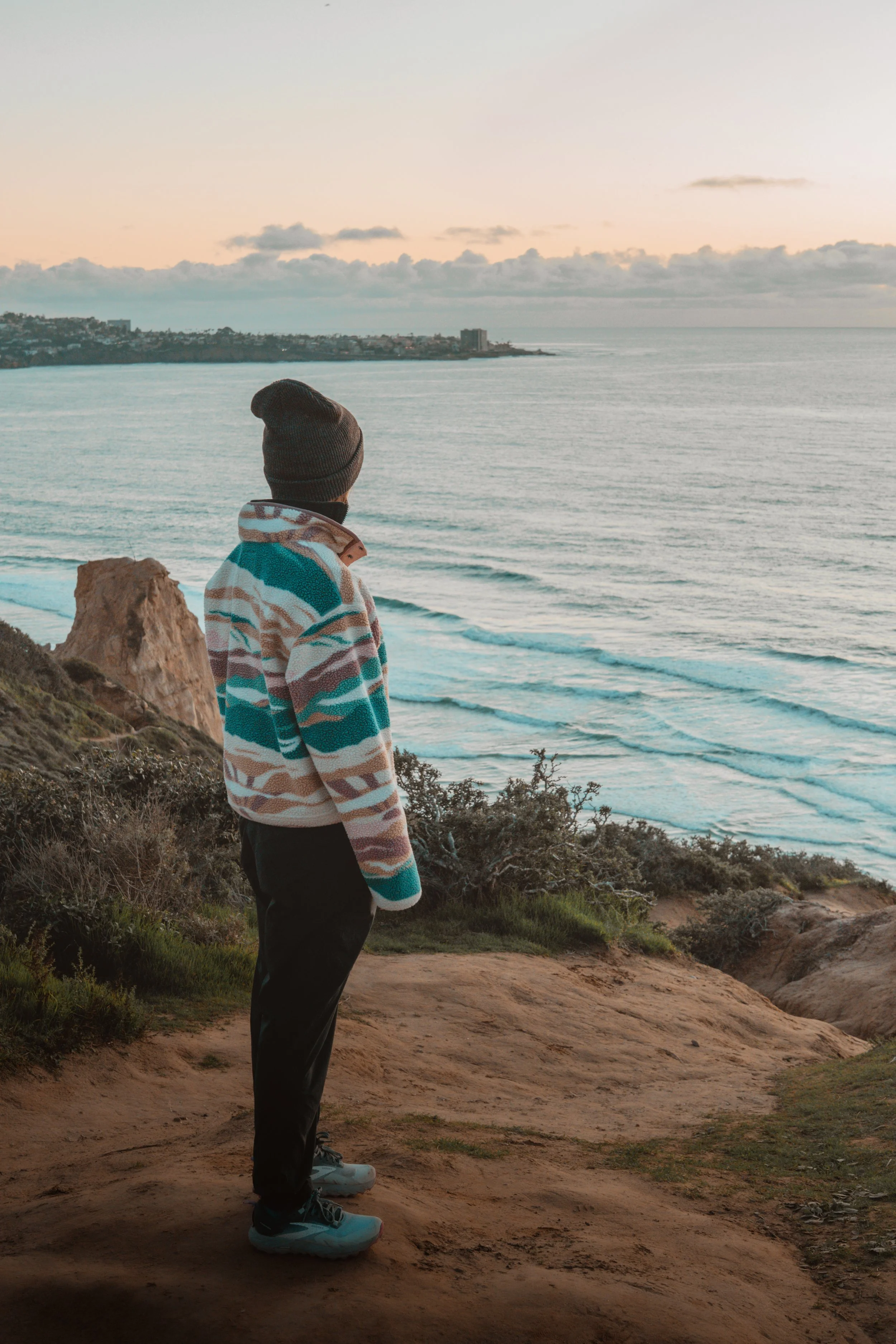 A person standing on a dirt path overlooking the ocean at sunset, wearing a colorful striped jacket, black pants, sneakers, and a beanie, with a coastal landscape and cliffs in the background.