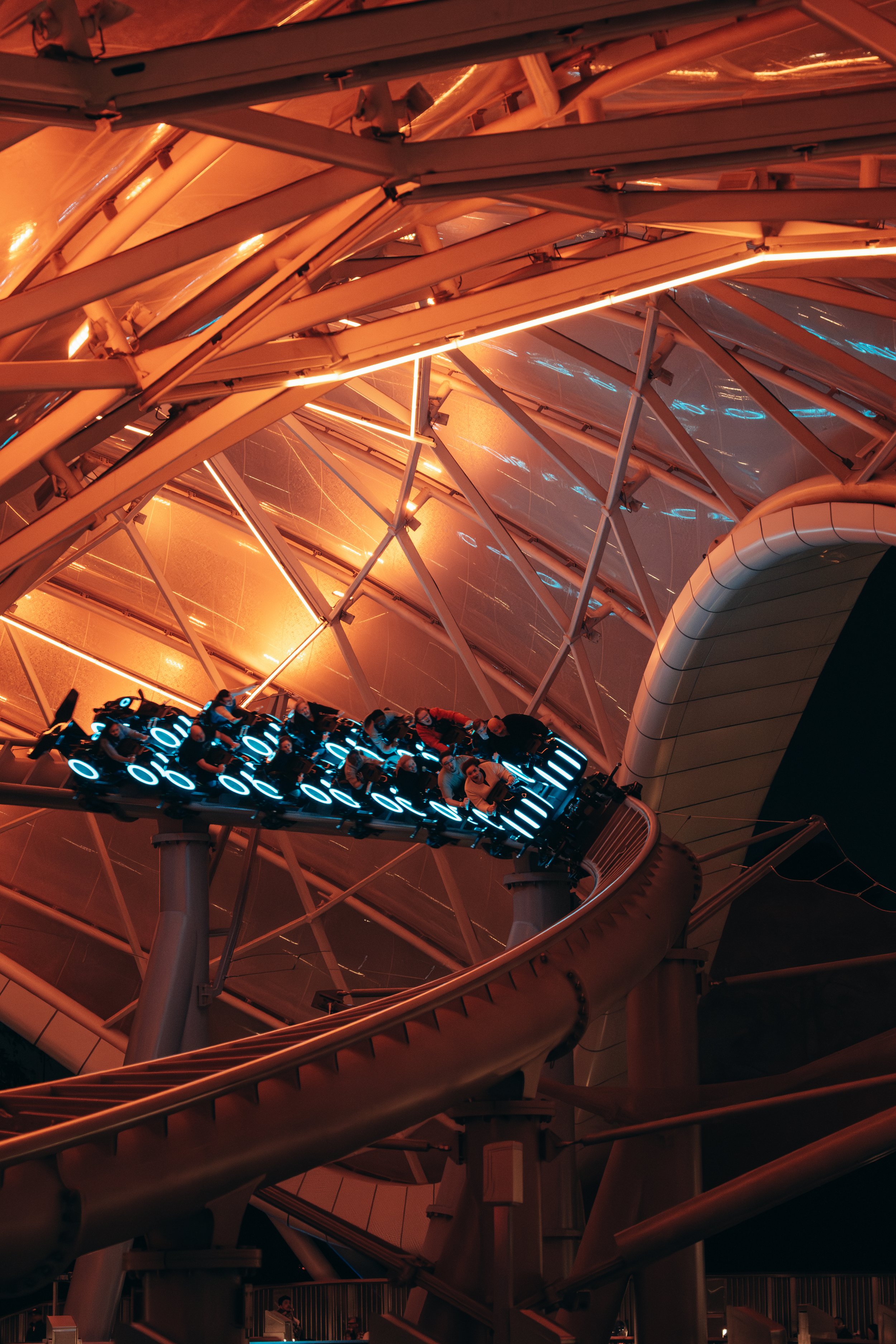 People riding a futuristic roller coaster at night, with glowing blue lights, inside a modern architectural structure with steel beams and glass panels illuminated with orange lighting.