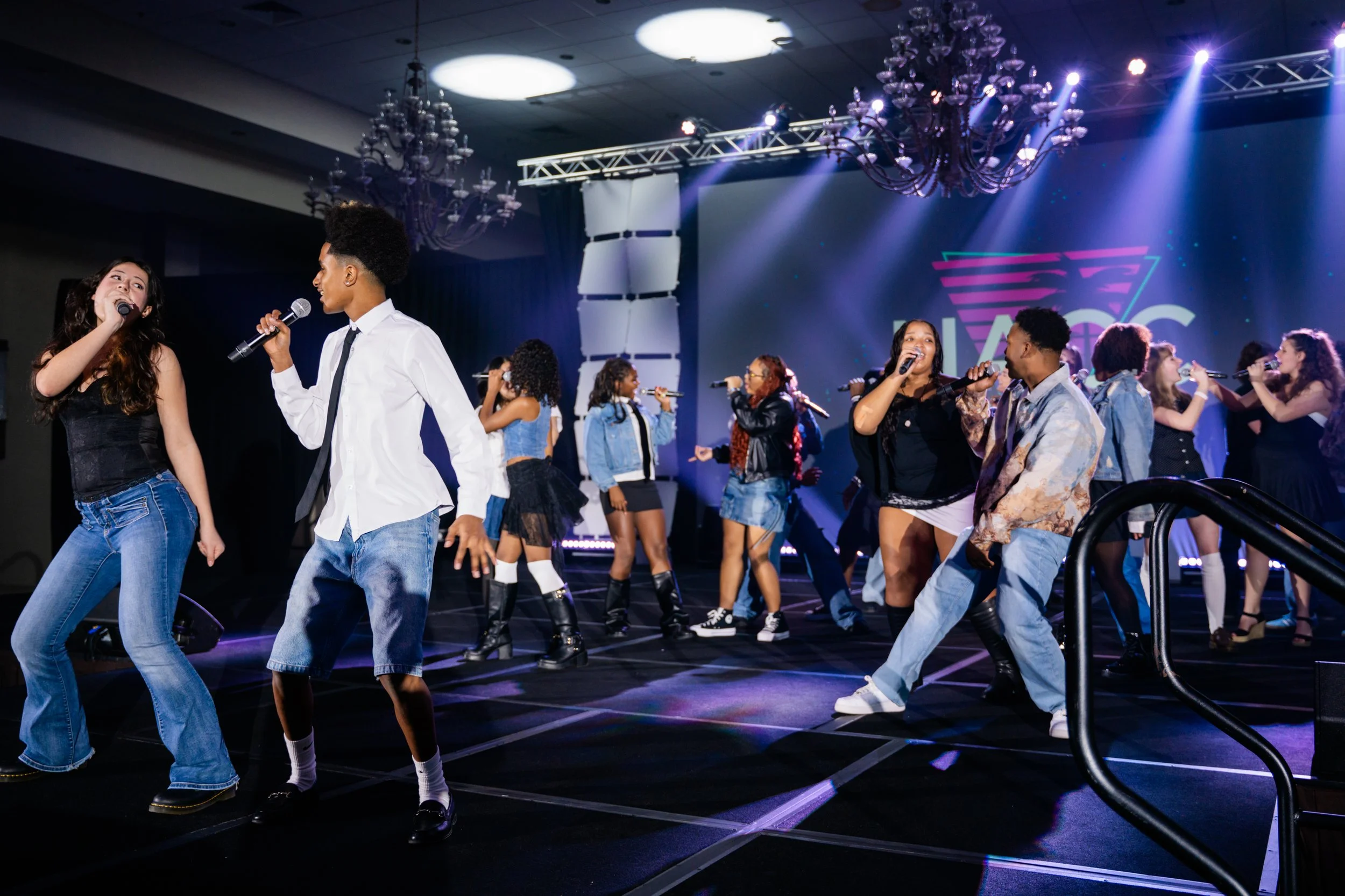 People singing and dancing on a stage with colorful lights and a large screen in the background.