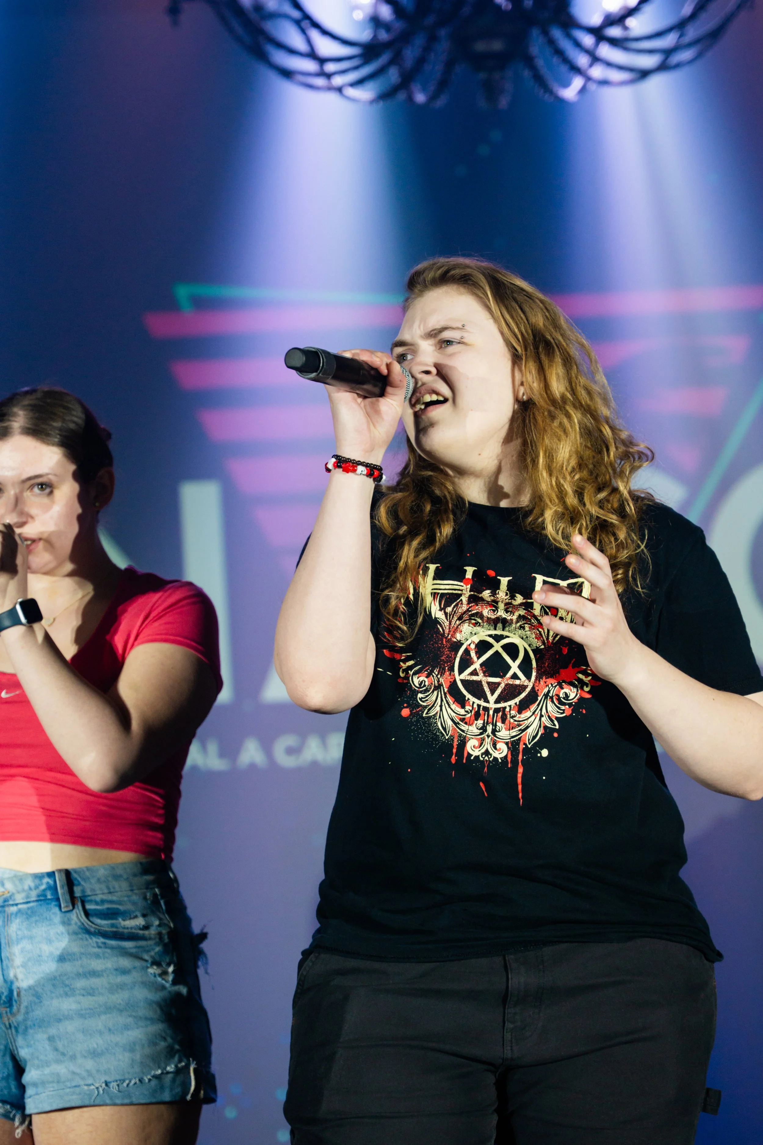 Two women performing on stage, one singing into a microphone, with colorful background lights.