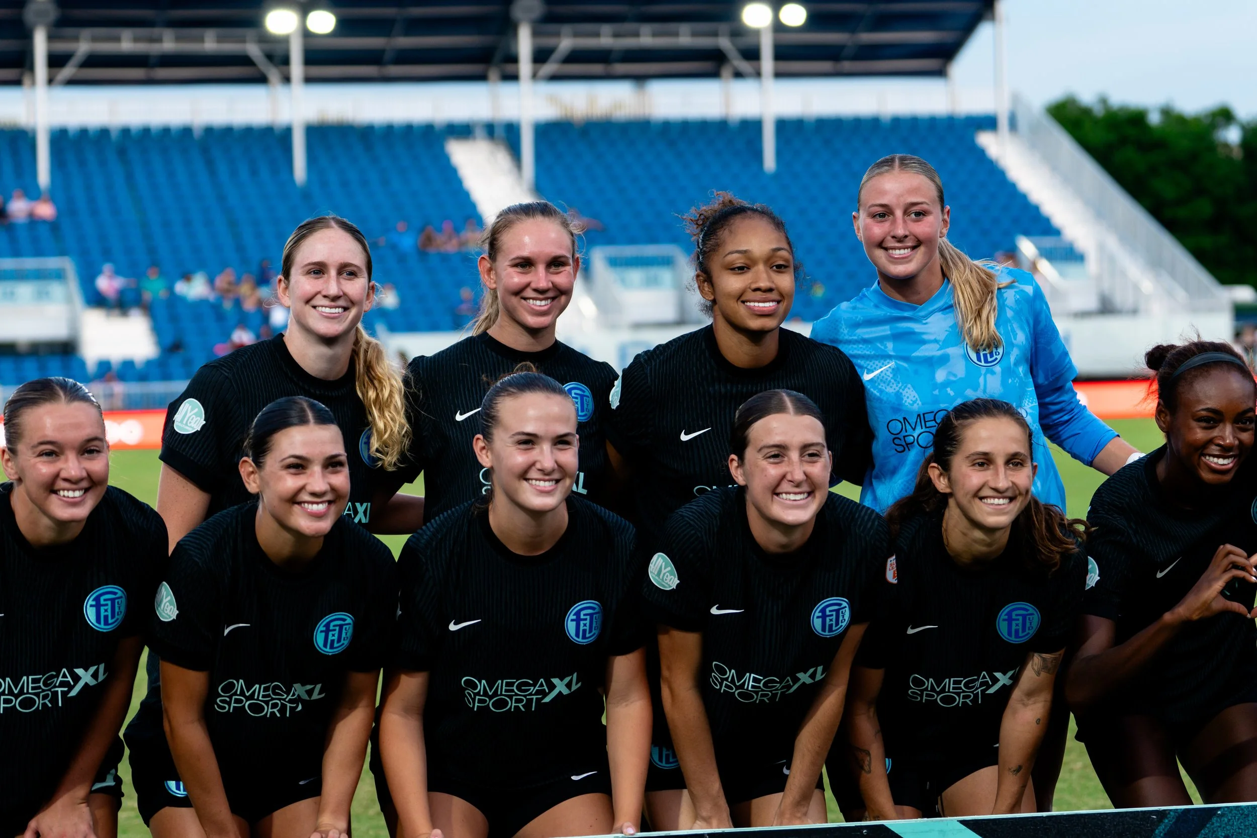 A group of female soccer players, wearing black and blue jerseys, smiling on a soccer field with a stadium and other players in the background.