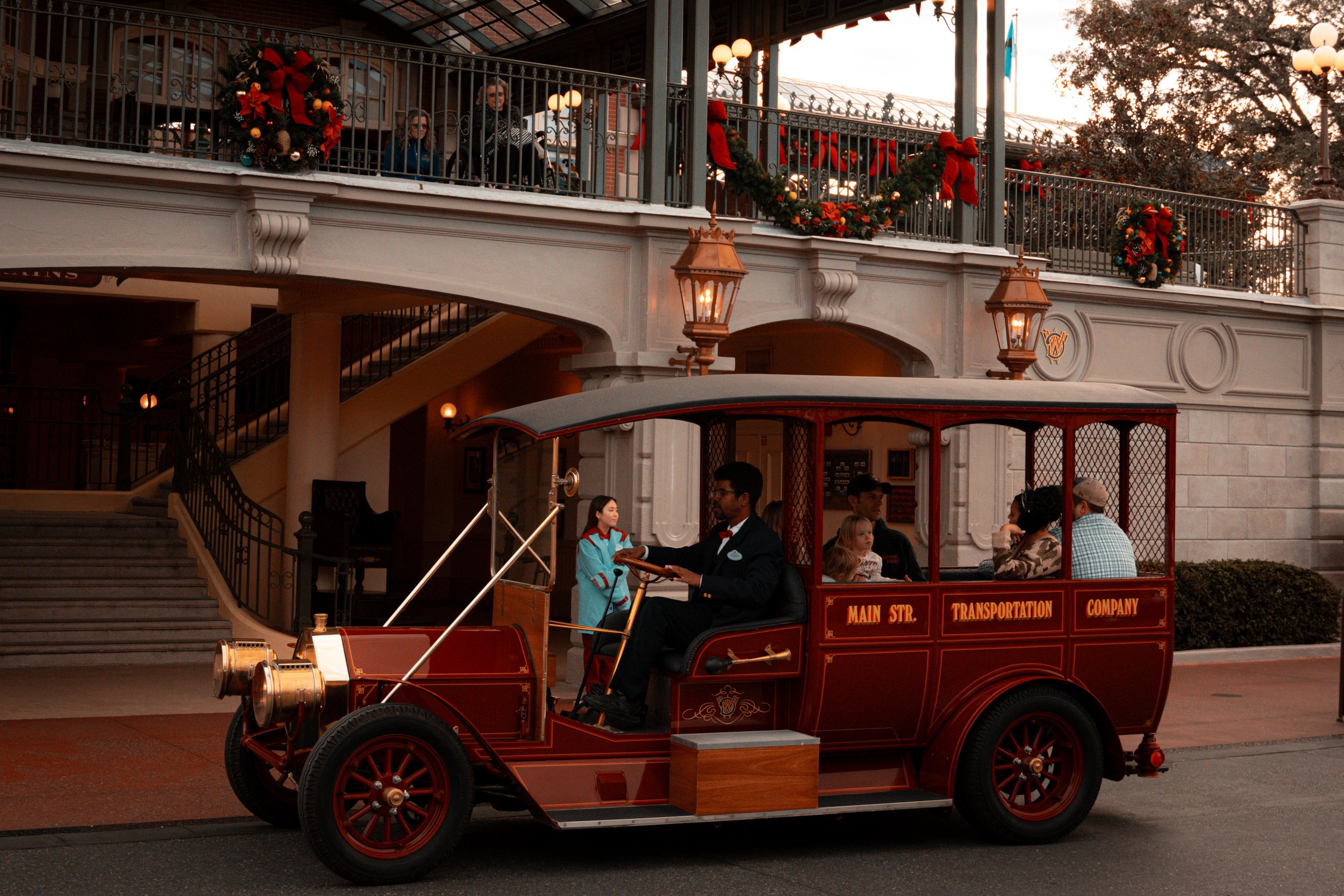 A vintage red trolley bus with gold details and the words 'Main Str. Transportation Company' on the side, driving on a street in front of a building decorated for Christmas with wreaths and bows. The trolley bus is carrying passengers and is driven b