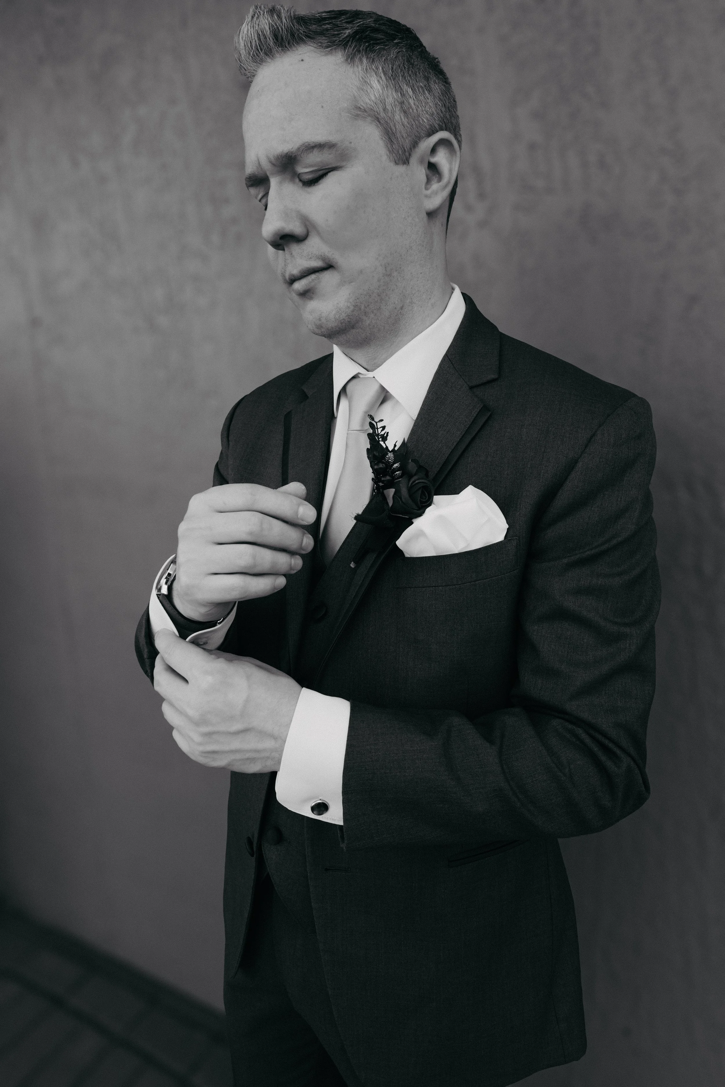 A man in a suit adjusting his cufflink, with a boutonniere on his lapel and a pocket square, standing against a plain wall.