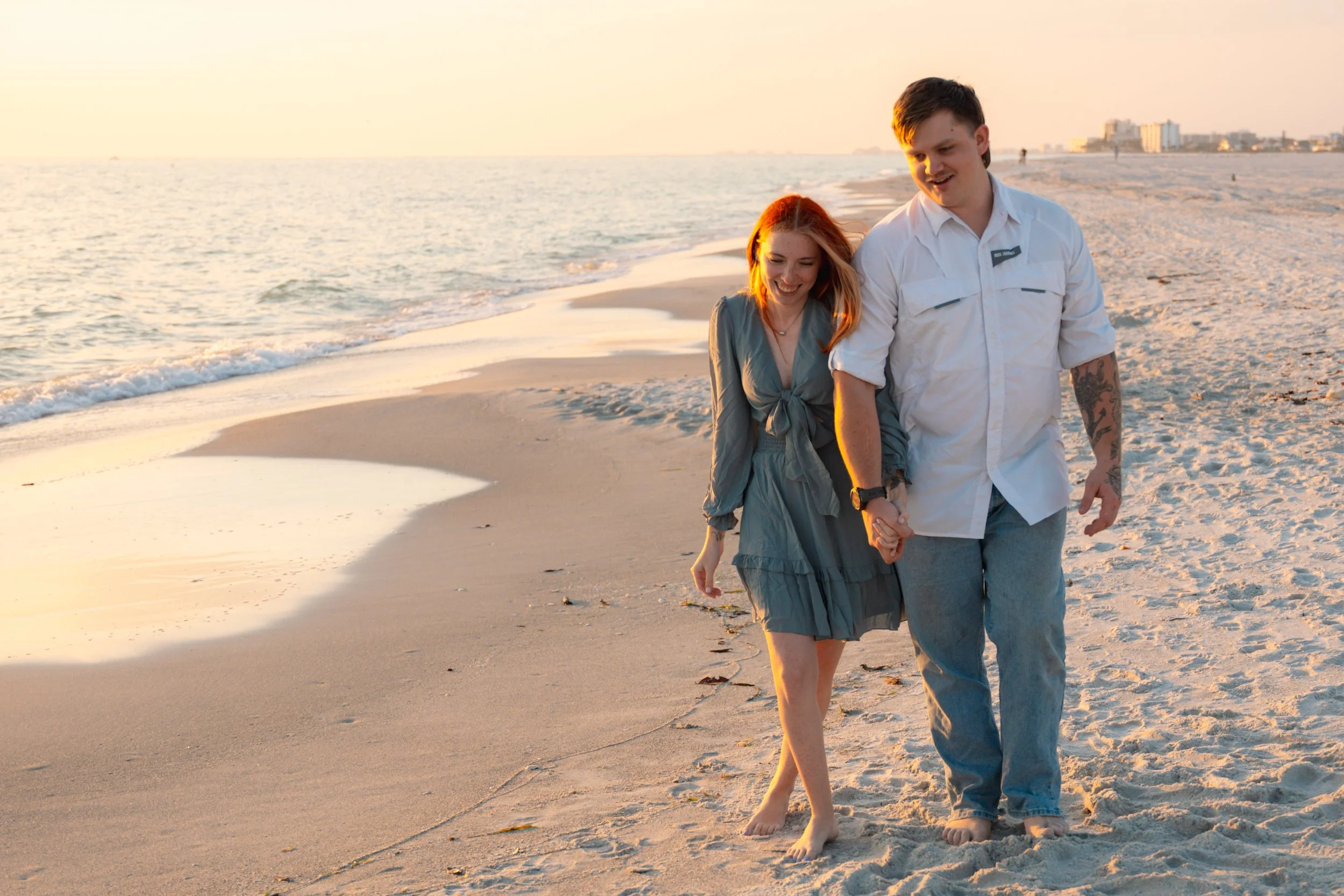 A happy couple walking on the beach during sunset, holding hands and smiling.