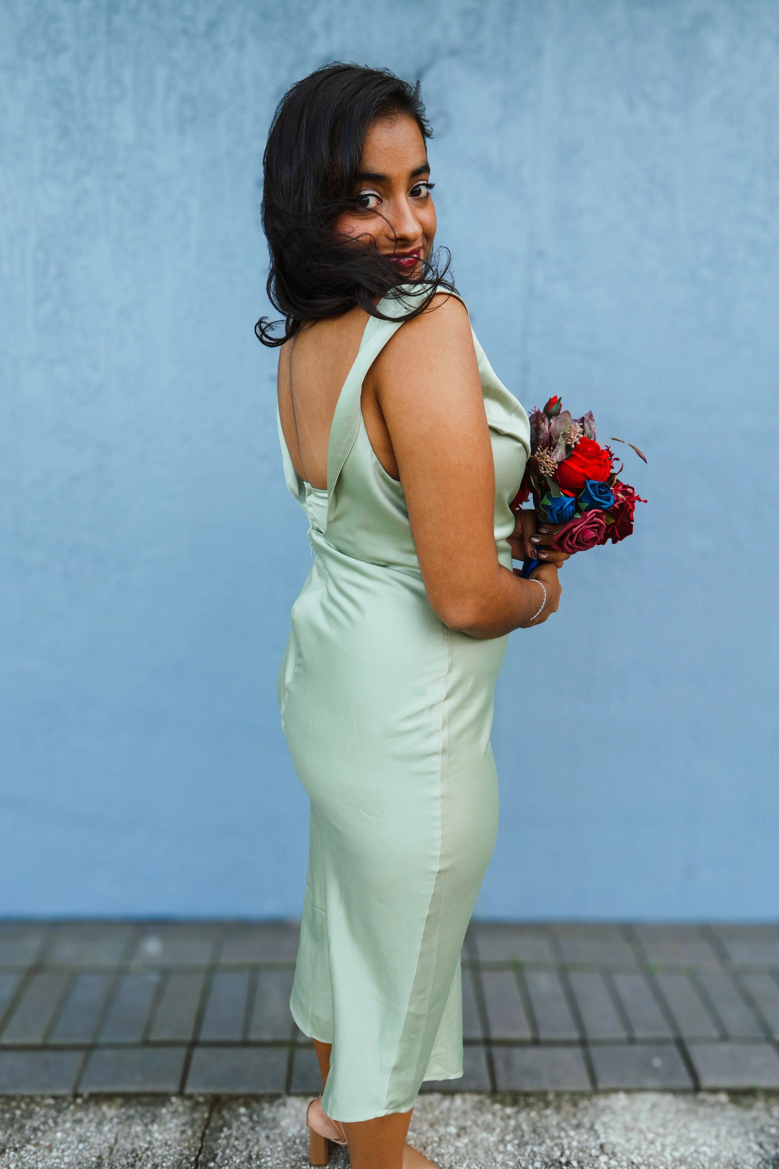 Woman in a light green dress holding a bouquet of red, blue, and purple roses, standing outdoors against a blue wall.