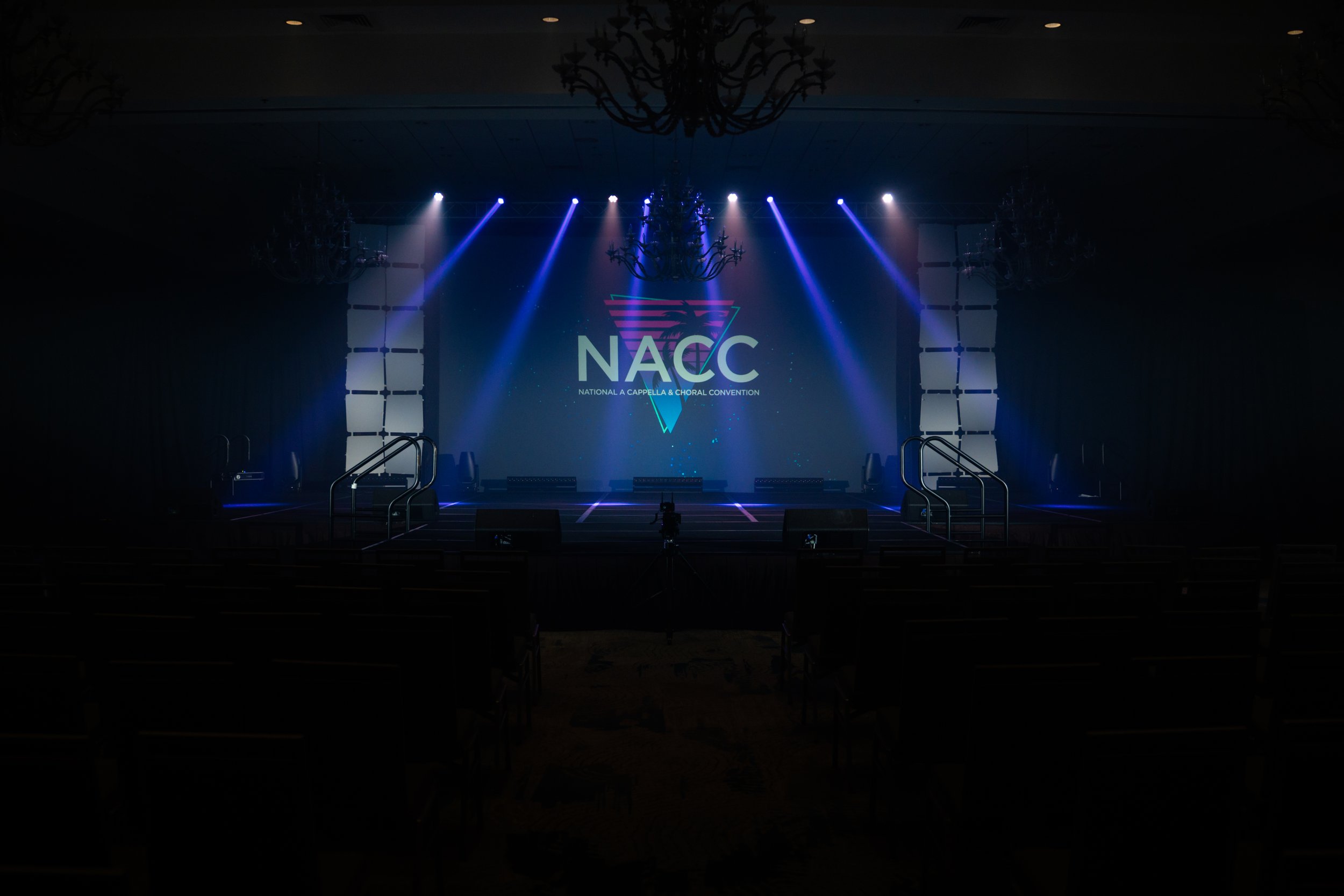 Stage set up for the National A Cappella & Choral Convention with lighting, sound equipment, and a large screen displaying the NACCC logo.