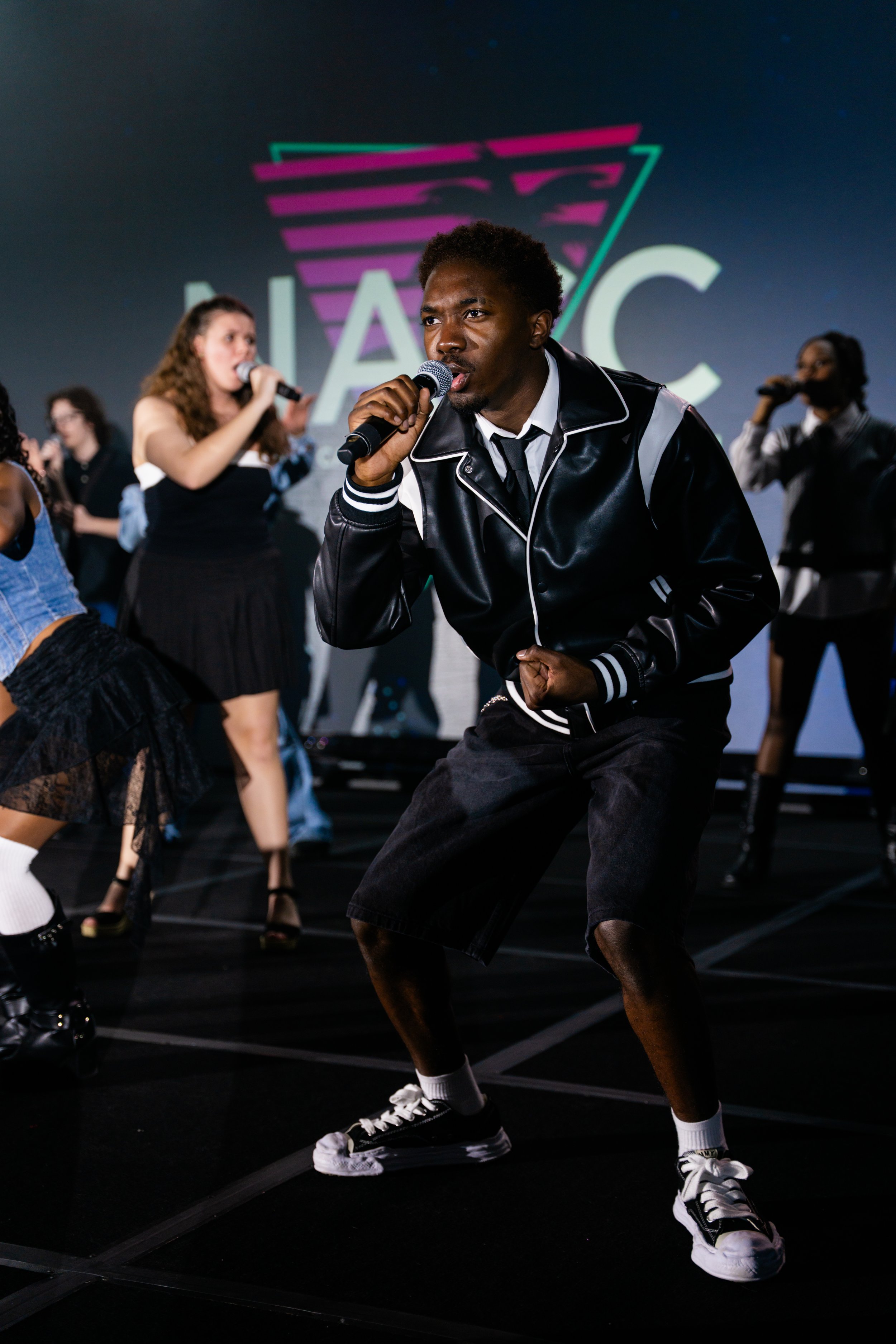 Young African American man singing into a microphone on stage, surrounded by women dancing, with a large logo in the background.
