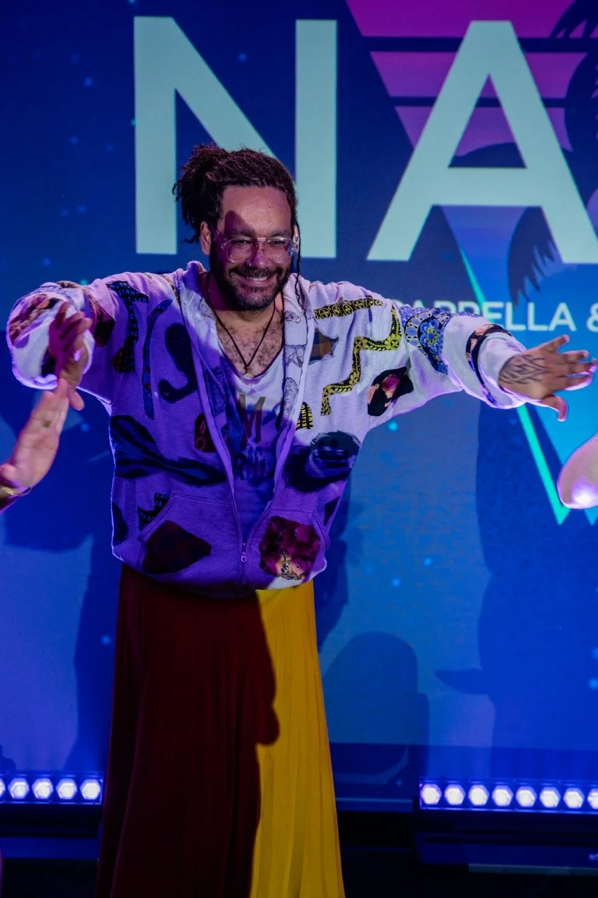 A man with dreadlocks and glasses dancing on stage with a big smile, wearing a colorful jacket and a yellow skirt, in front of a screen with the letters 'NA' visible.