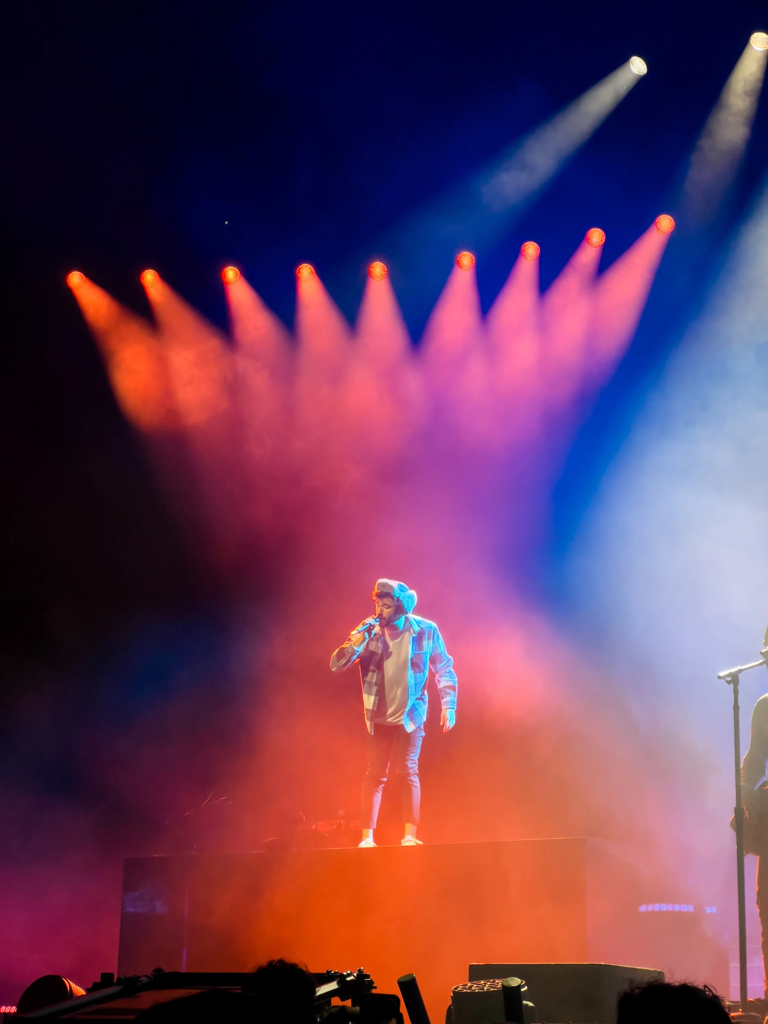 A singer on stage under colorful stage lights, wearing a plaid shirt and headphones, holding a microphone.