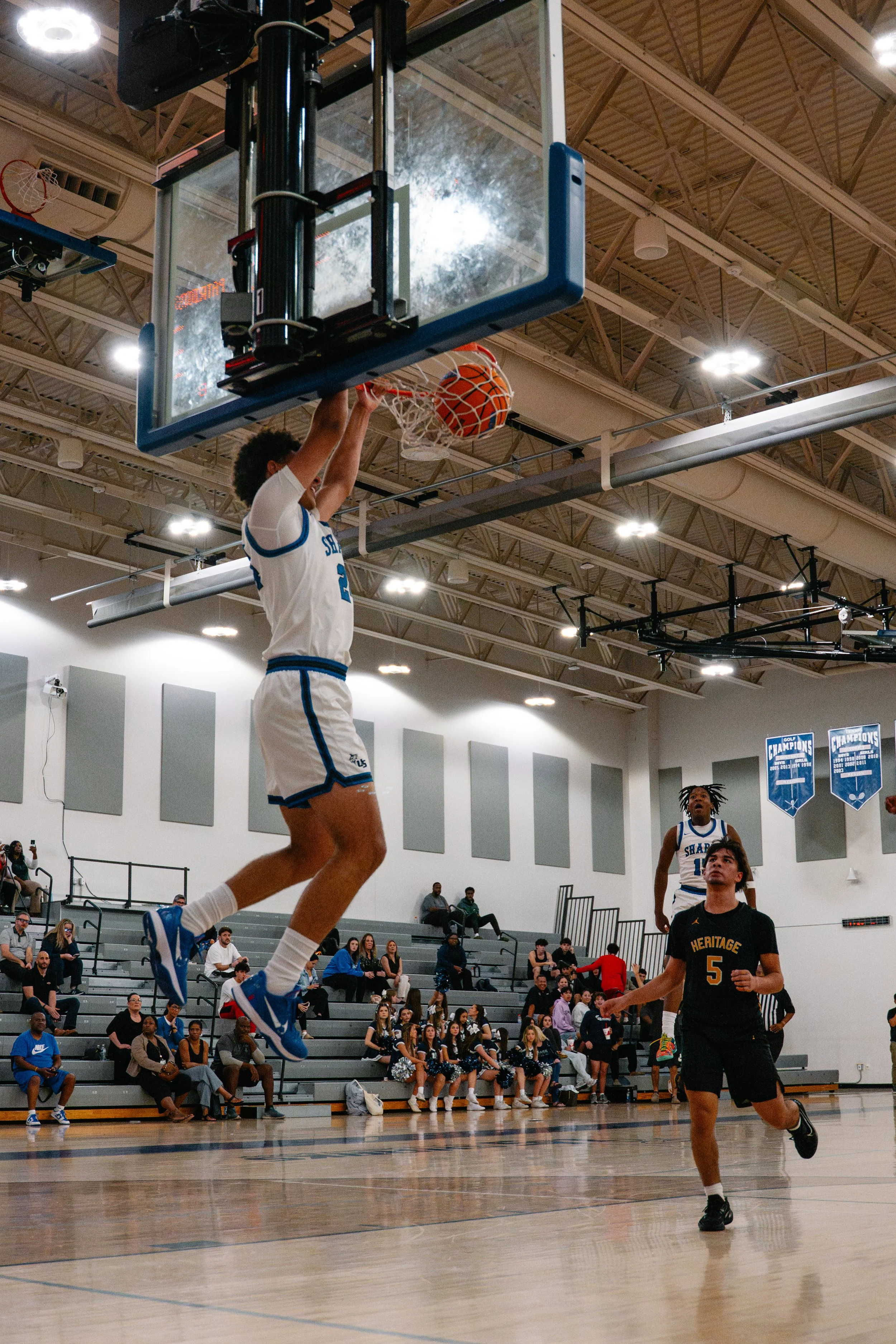 A basketball player in a white and blue jersey is jumping to make a dunk, with the basketball going through the hoop. Another player in a black jersey with the number 5 is running underneath, while spectators sit on bleachers in the background.