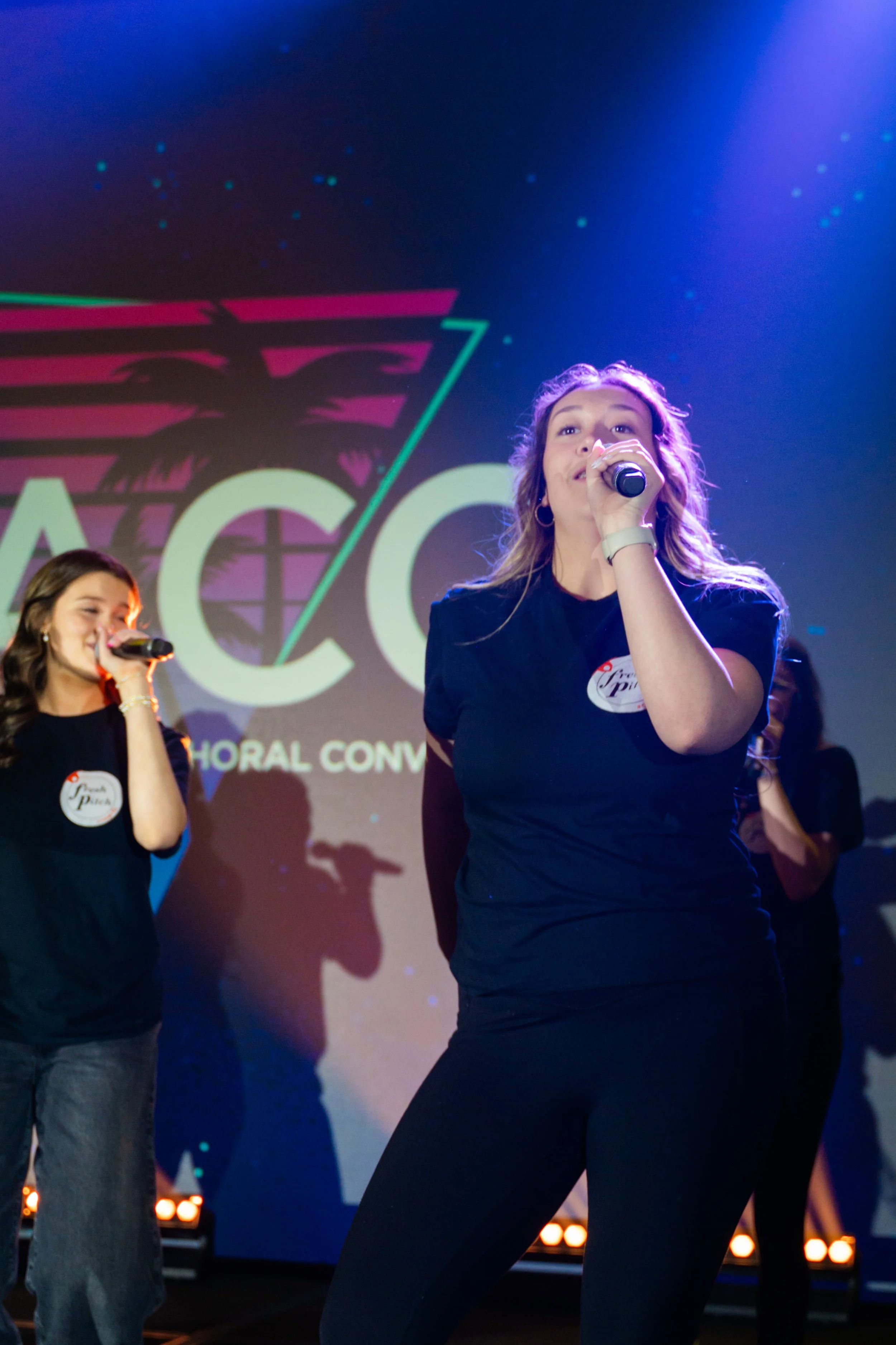 Two women singing on stage with microphones, one in the foreground and one in the background, under colorful stage lighting with a large screen displaying part of a logo.