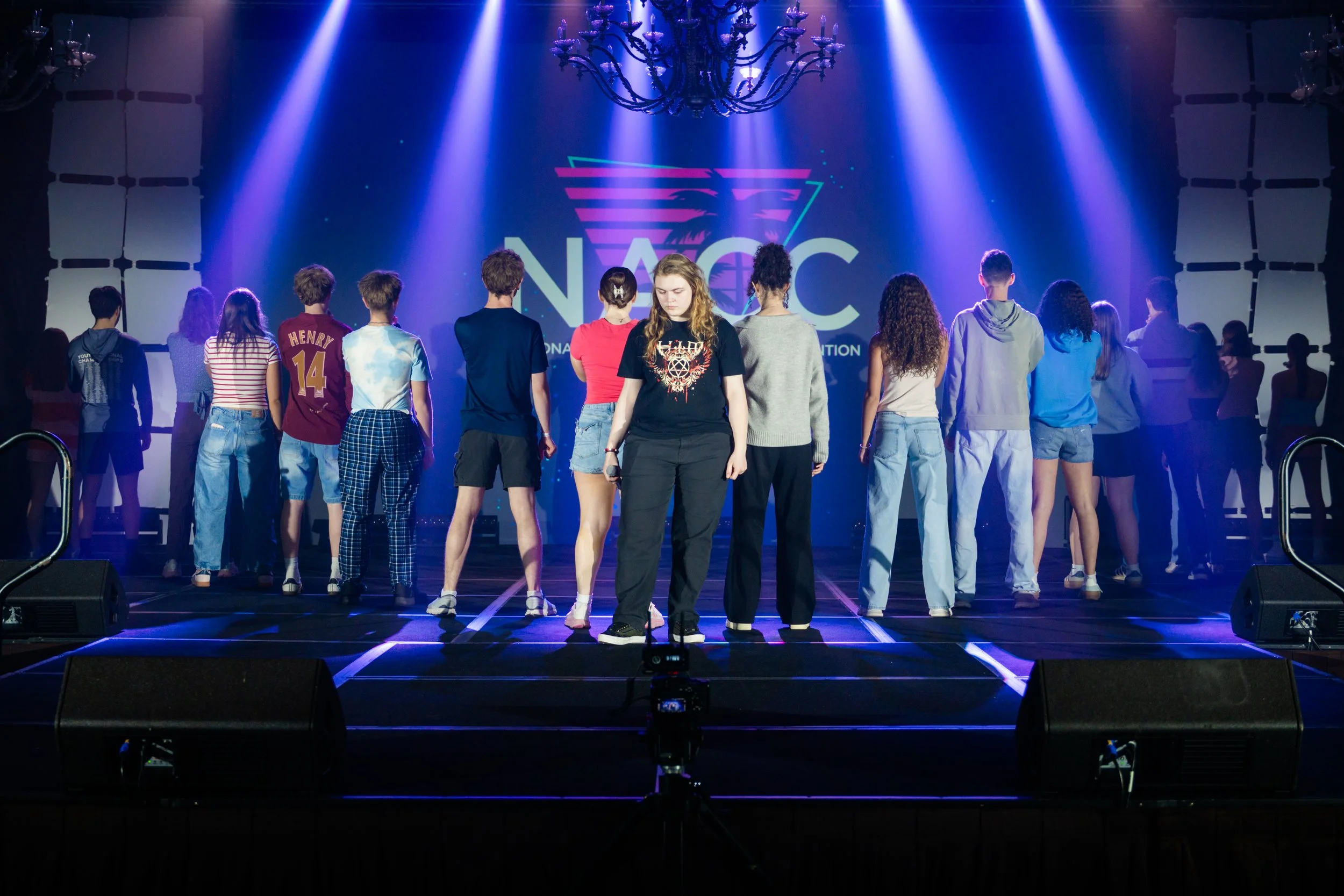 A group of young people standing on stage in front of a large screen displaying the NVC logo, illuminated by colorful stage lights.