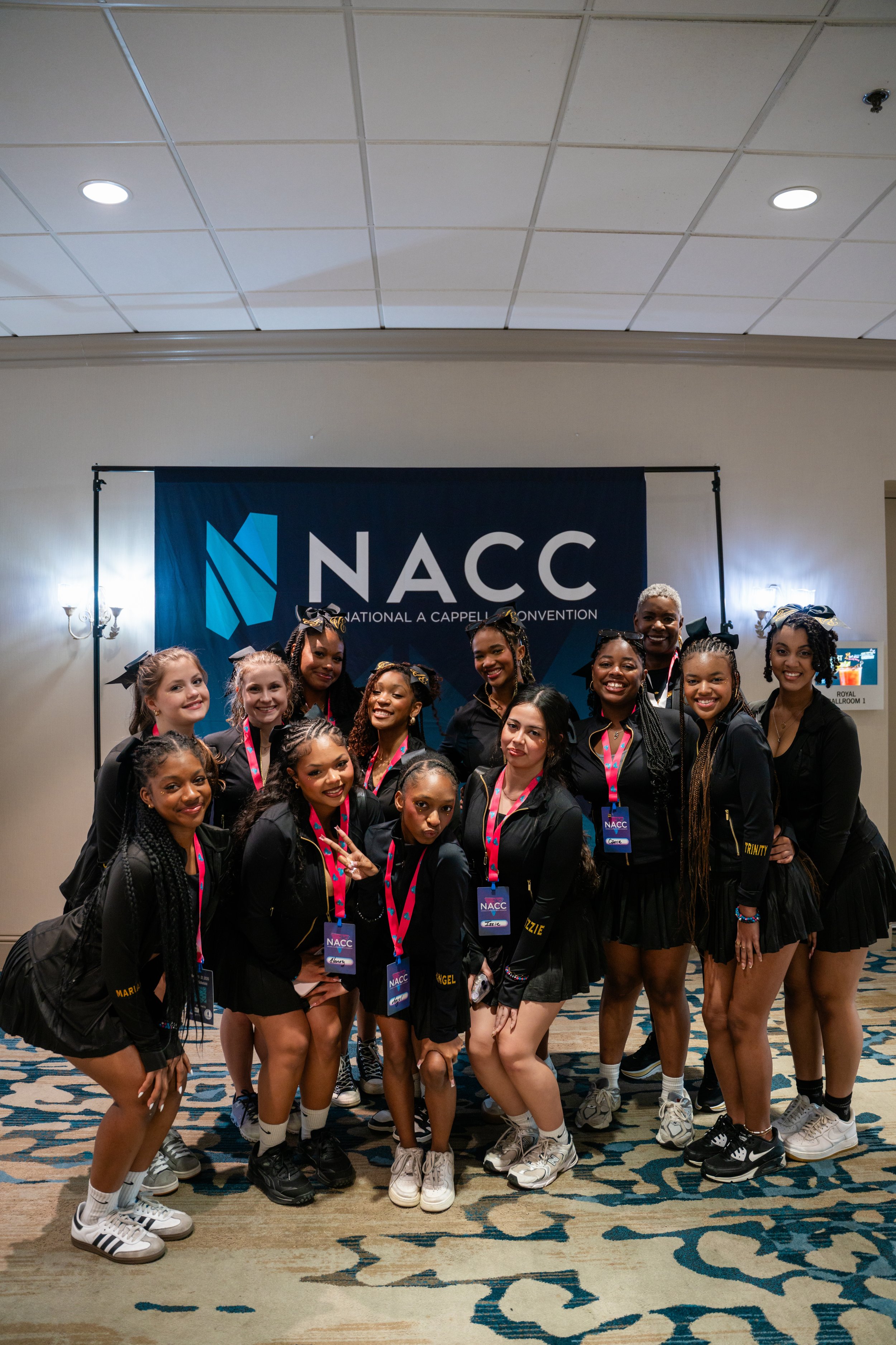 Group of cheerleaders posing in front of a banner that reads 'NACC National A Cappella Convention', indoors with patterned carpet and ceiling lights.