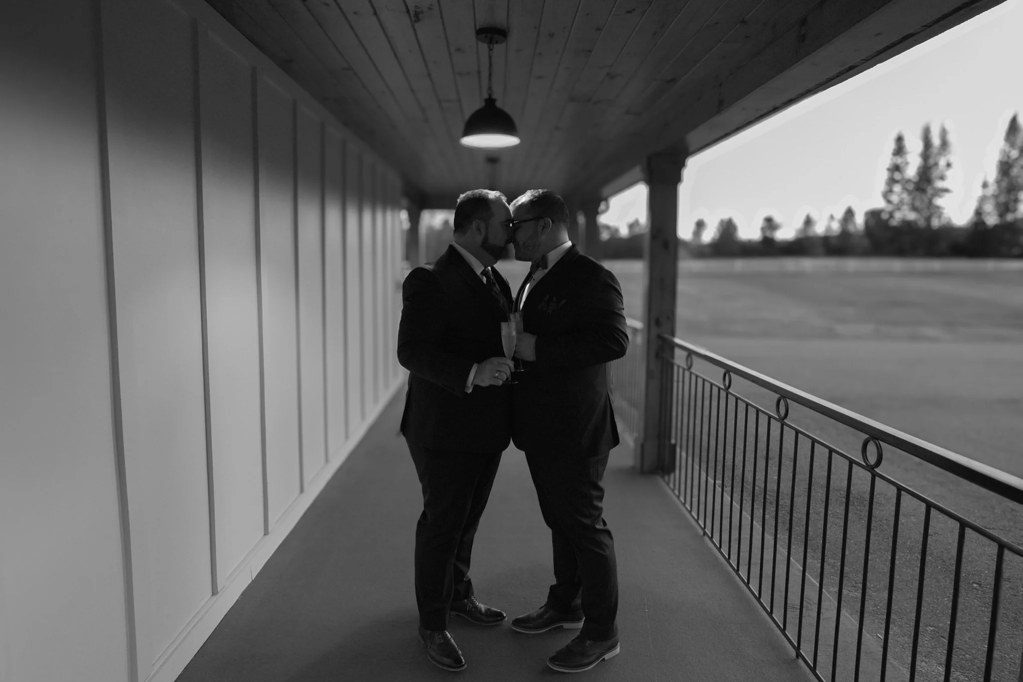 Two men dressed in suits are standing close, touching foreheads, sharing a tender moment on a covered balcony overlooking an open field with trees in the background.