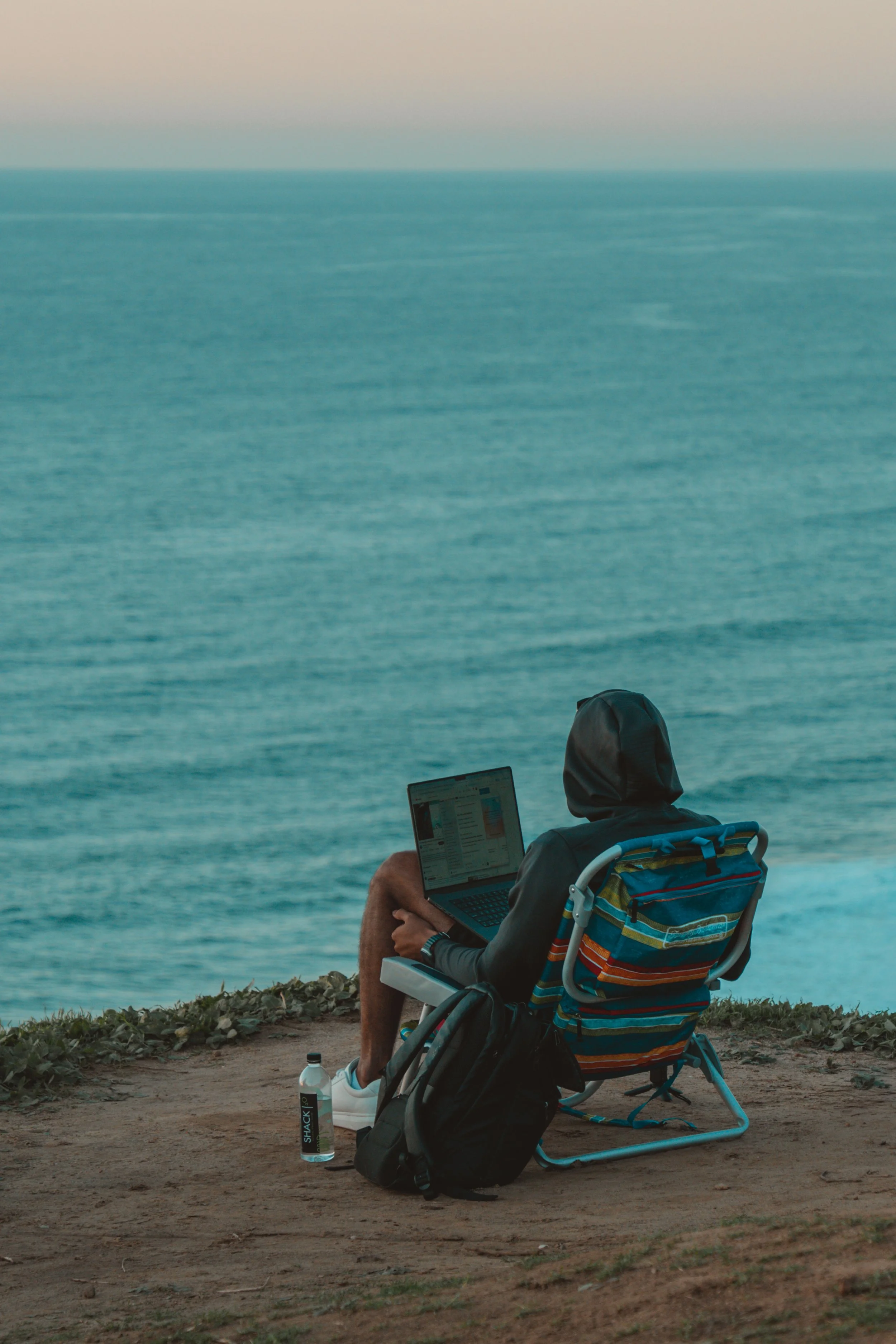 Person sitting in a foldable chair on a grassy cliff overlooking the ocean, working on a laptop. A water bottle and backpack are next to them.