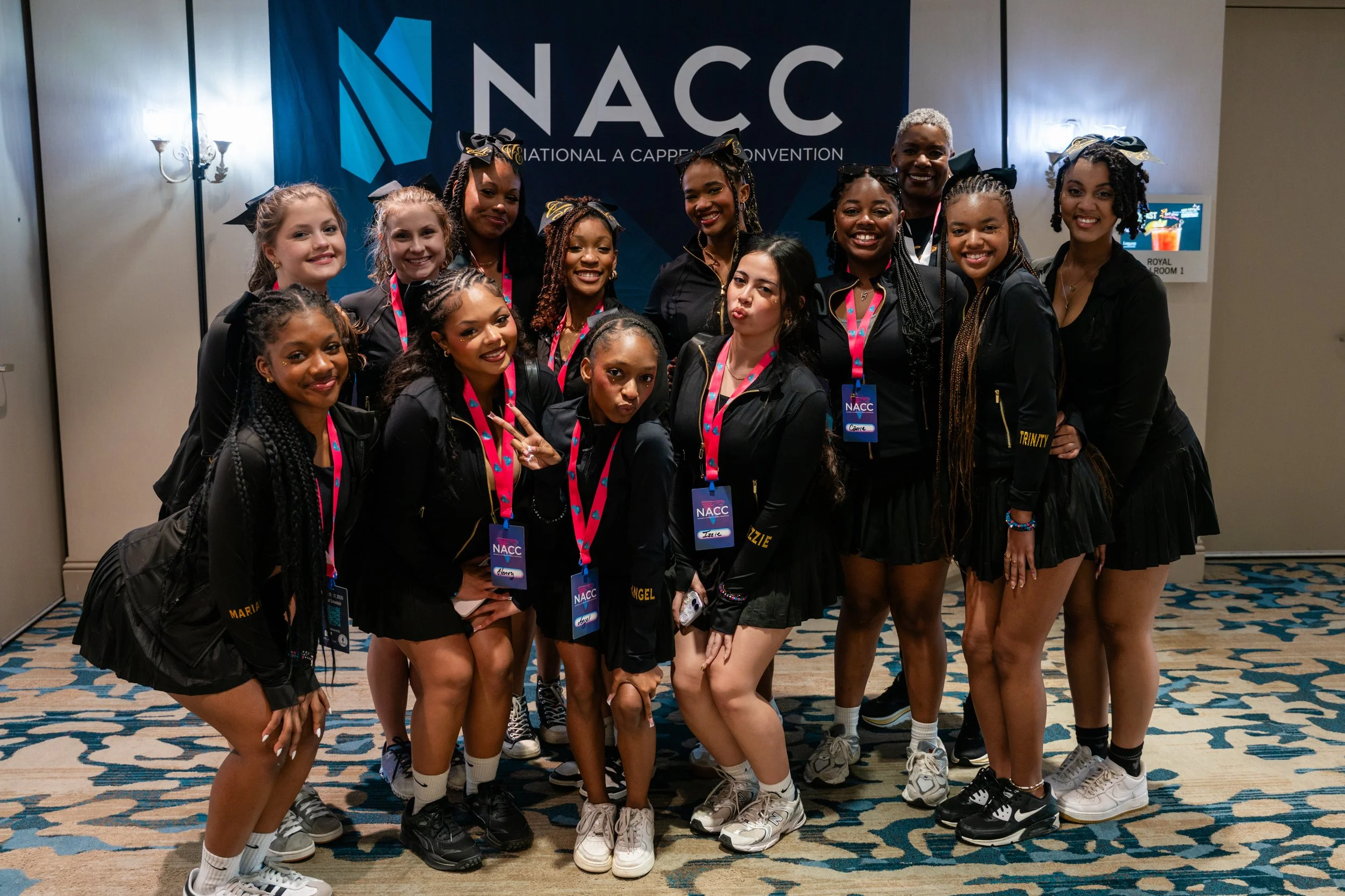 A group of young women posing together at the NAC conference, wearing black jackets and medals, with a large NAC sign in the background.