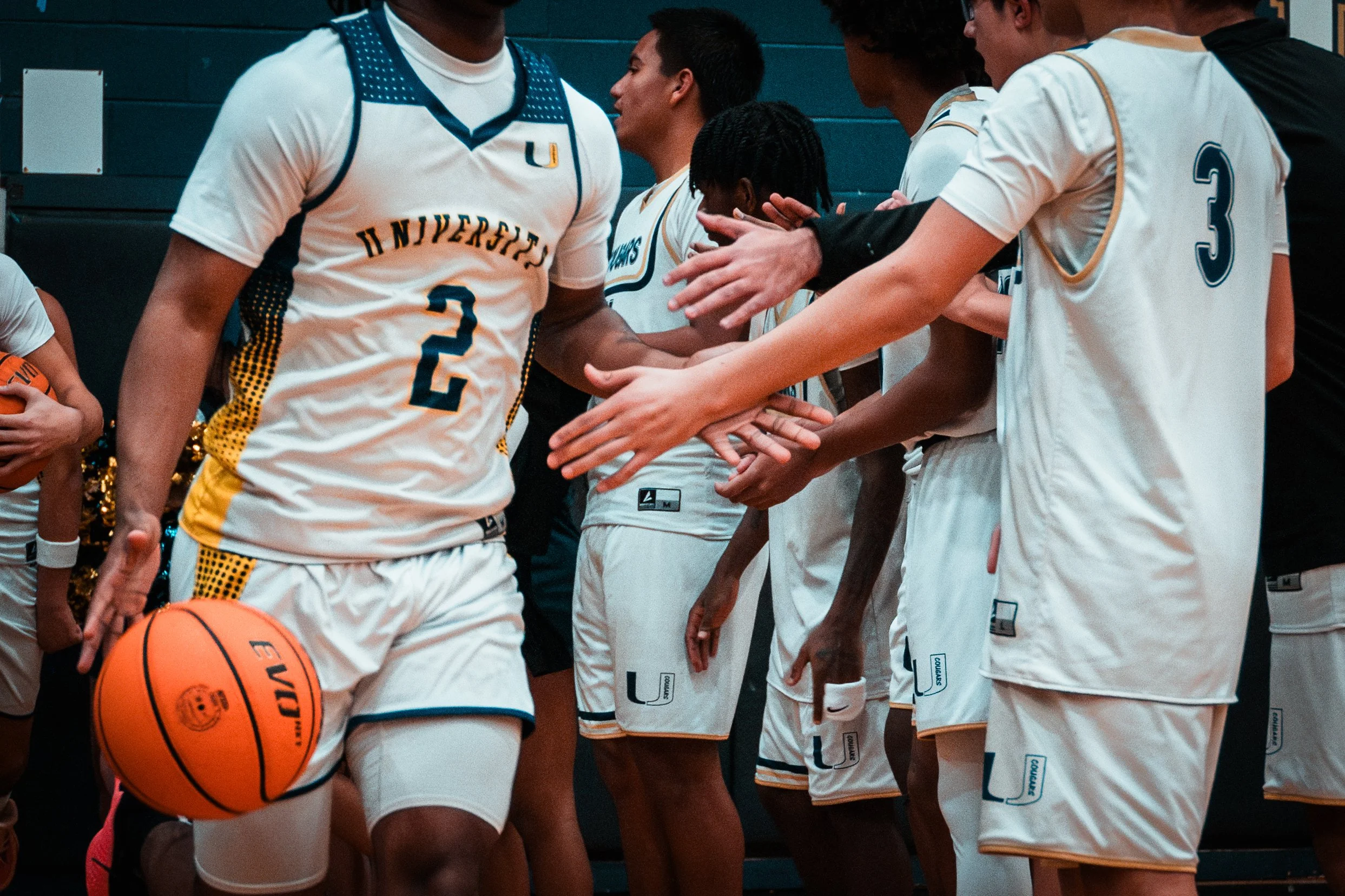 A group of basketball players in white uniforms with royal blue and gold accents, forming a line in a gymnasium. One player is holding an orange basketball, and they are engaging in a team huddle or high-five before a game or practice.
