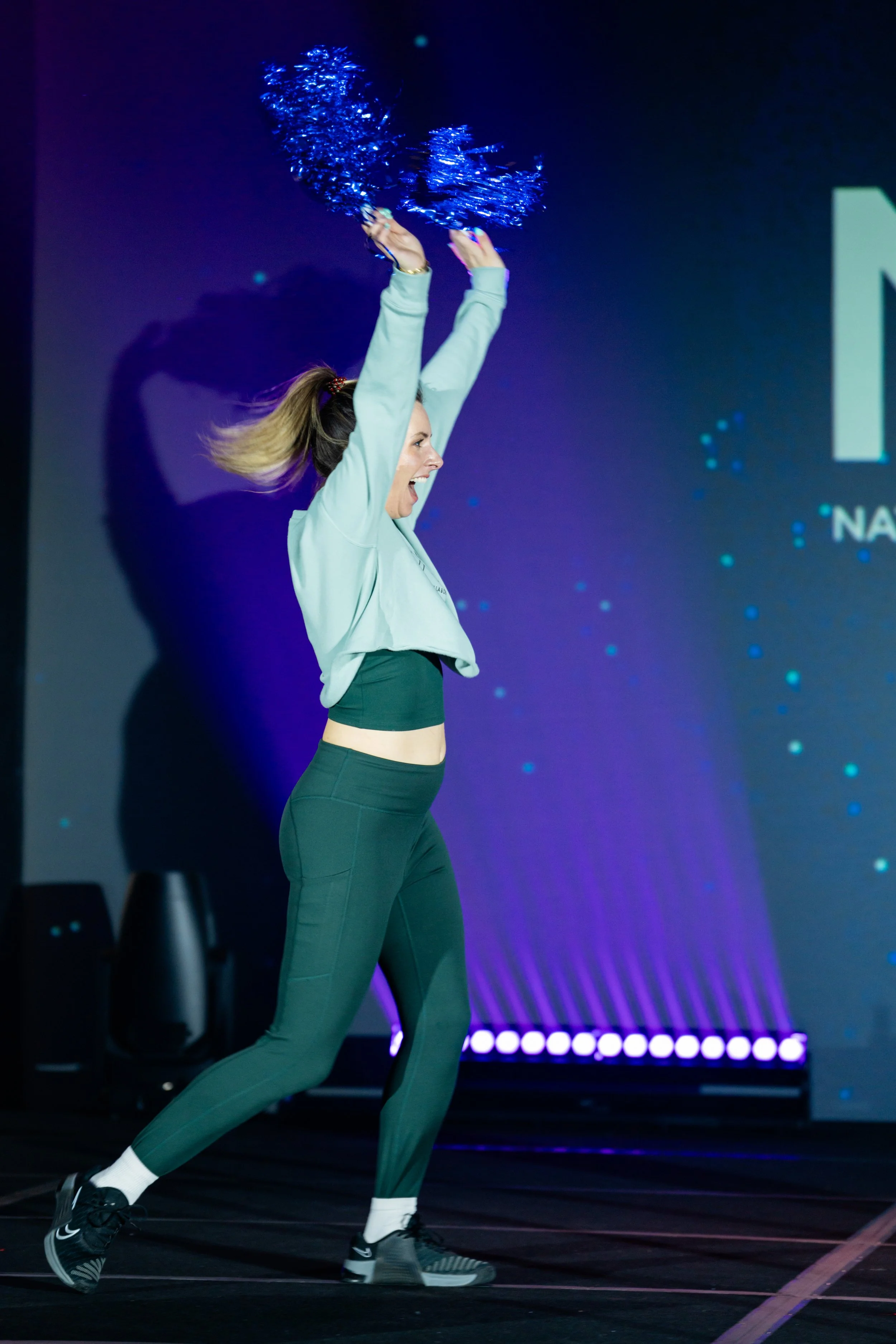 A woman in athletic clothing holding blue pom-poms and running or jumping on a stage with a dark background and colorful lights.