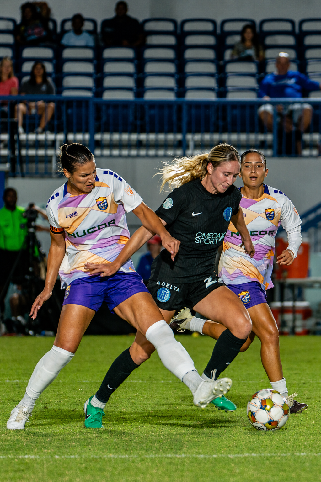Two female soccer players, one in a black uniform and two in white and purple uniforms, competing for the ball on the field during a match.