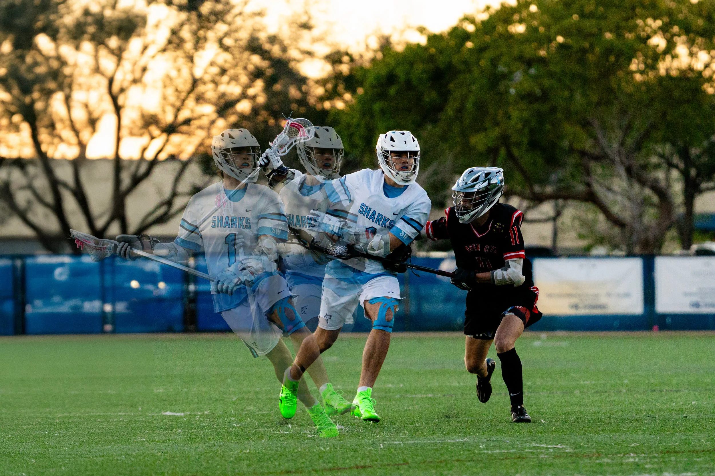 Lacrosse players in white and black uniforms competing on a green field at sunset.