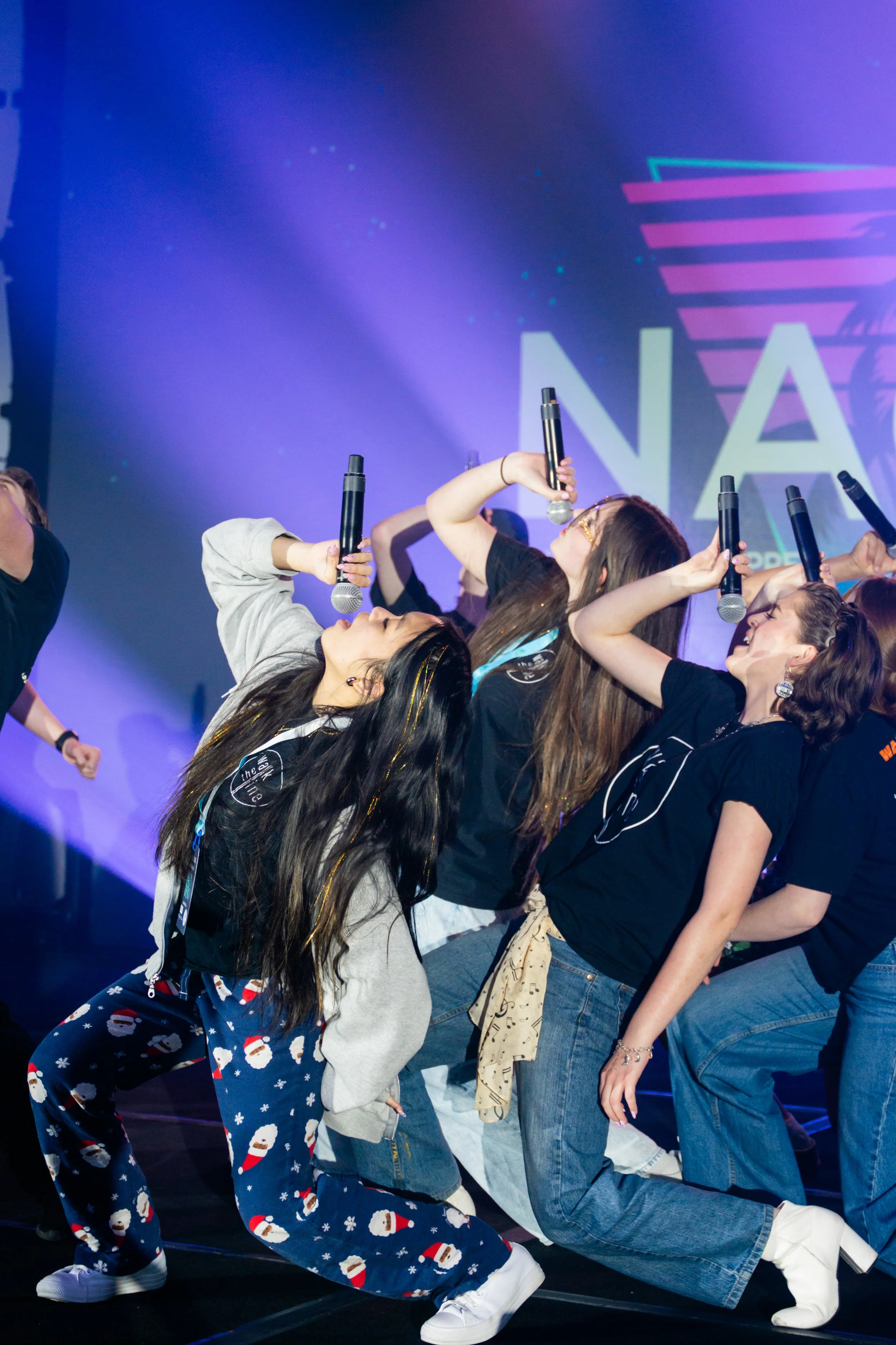Group of young women dancing on stage holding microphones, with colorful lights and a large sign that reads 'NA' in the background.