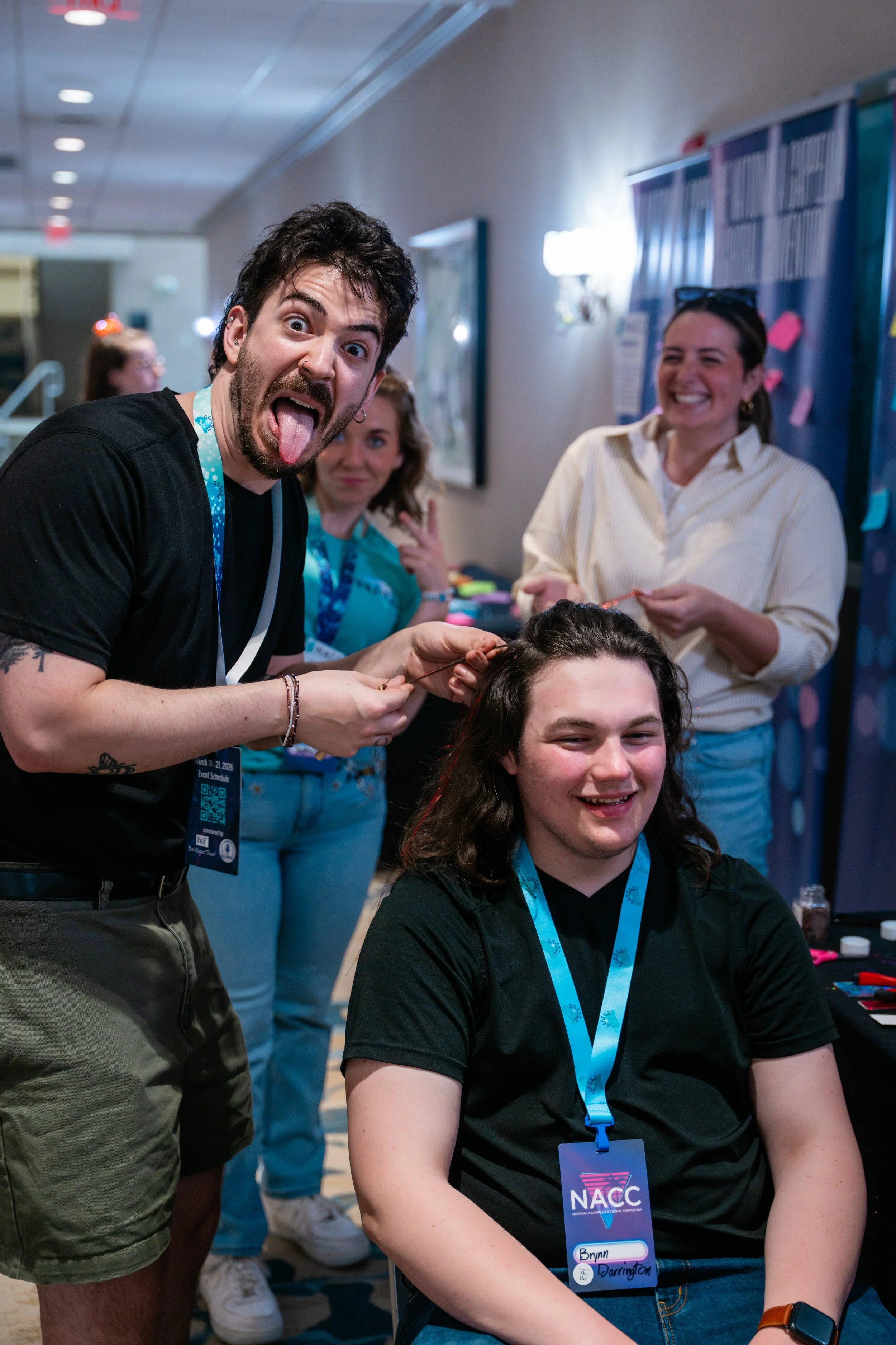 A group of people at a convention, with a man sticking out his tongue and making a funny face, while a woman with glasses and a big smile looks on. The woman has dark hair and is wearing a white shirt, and another woman with curly hair and a blue shi