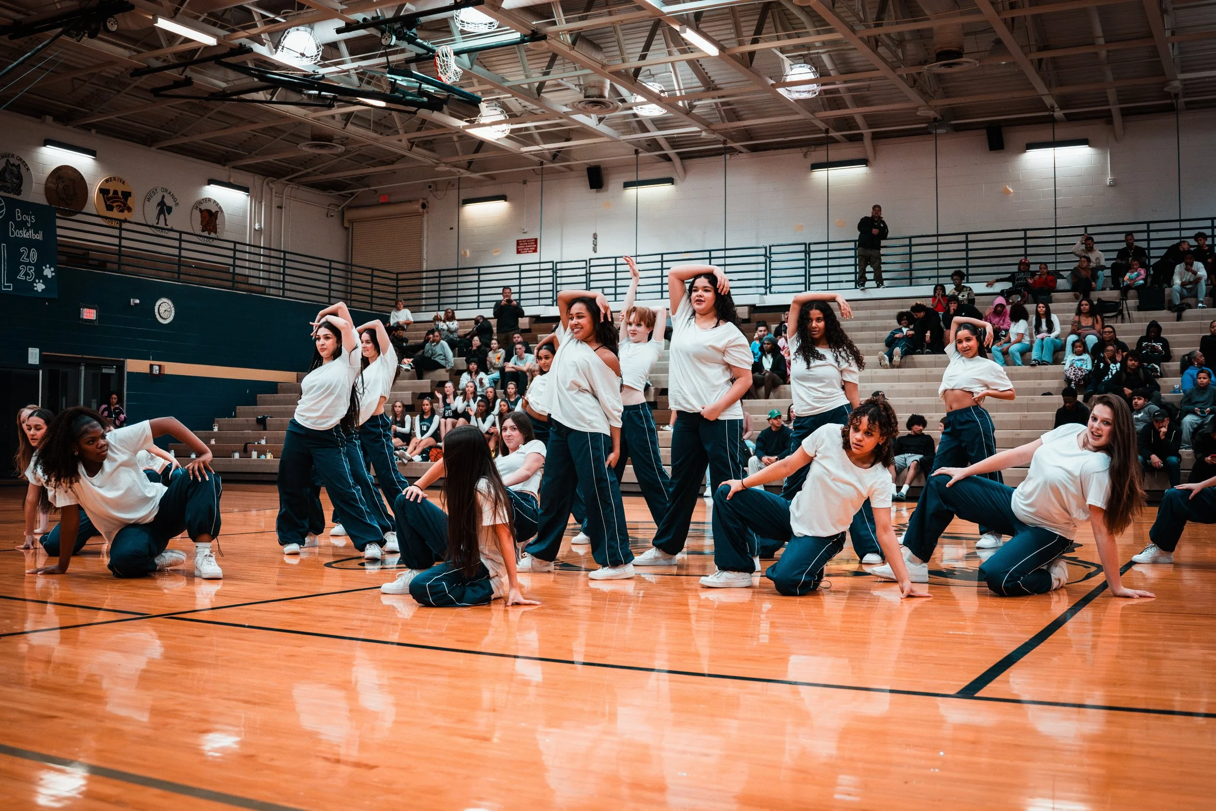 Group of young girls performing a dance routine in a gymnasium, with an audience seated on the bleachers watching.