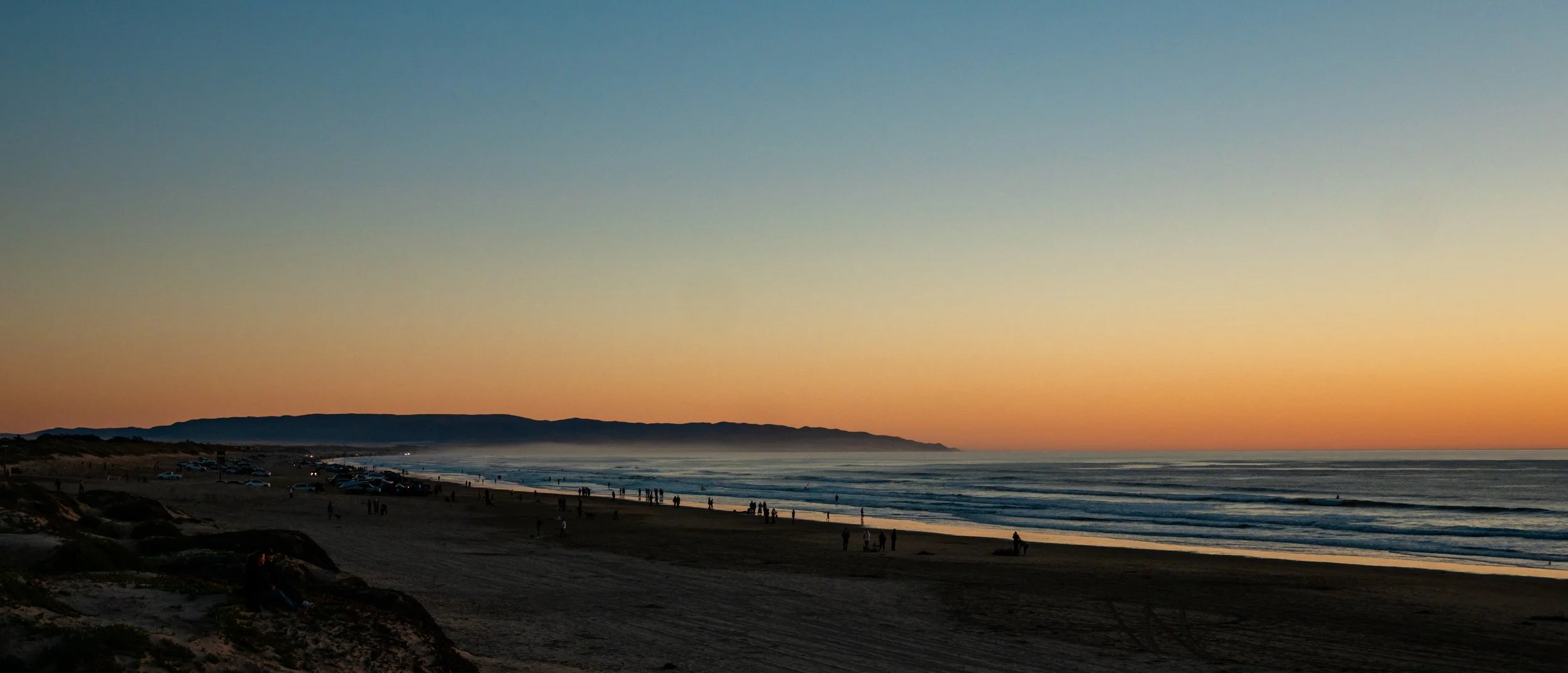 Sunset over a beach with people walking along the shore, parked cars, and a distant mountain range in the background.