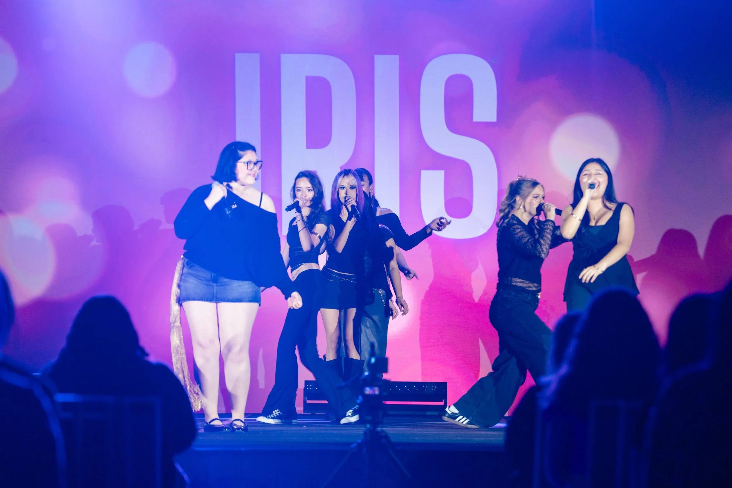 Five young women perform on stage with a pink and purple background displaying the words 'HBO'. They are singing into microphones, dressed in black outfits, with an audience visible in the foreground.