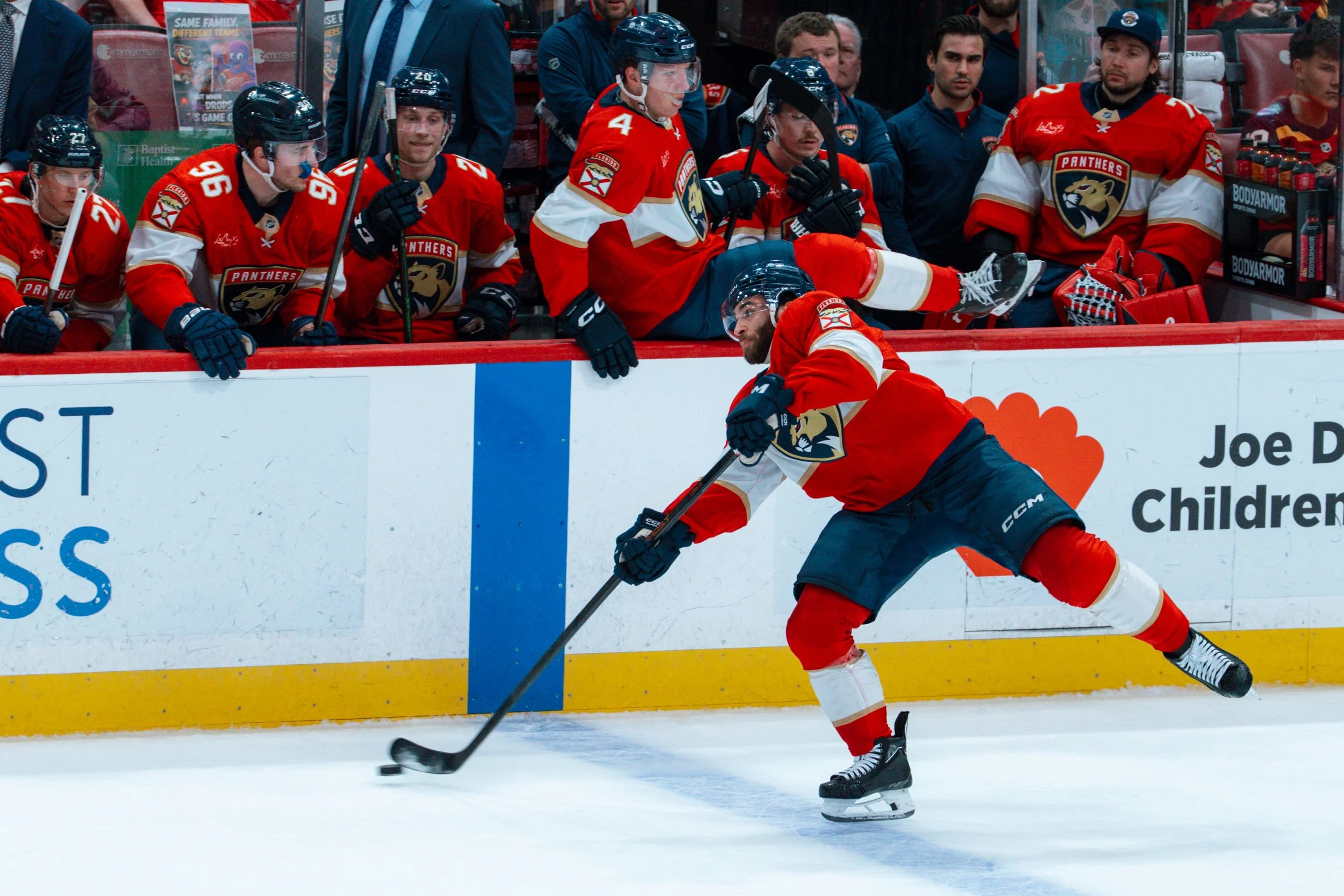 Hockey players on the ice during a game, with one player skating and handling the puck, while others sit on the bench watching.