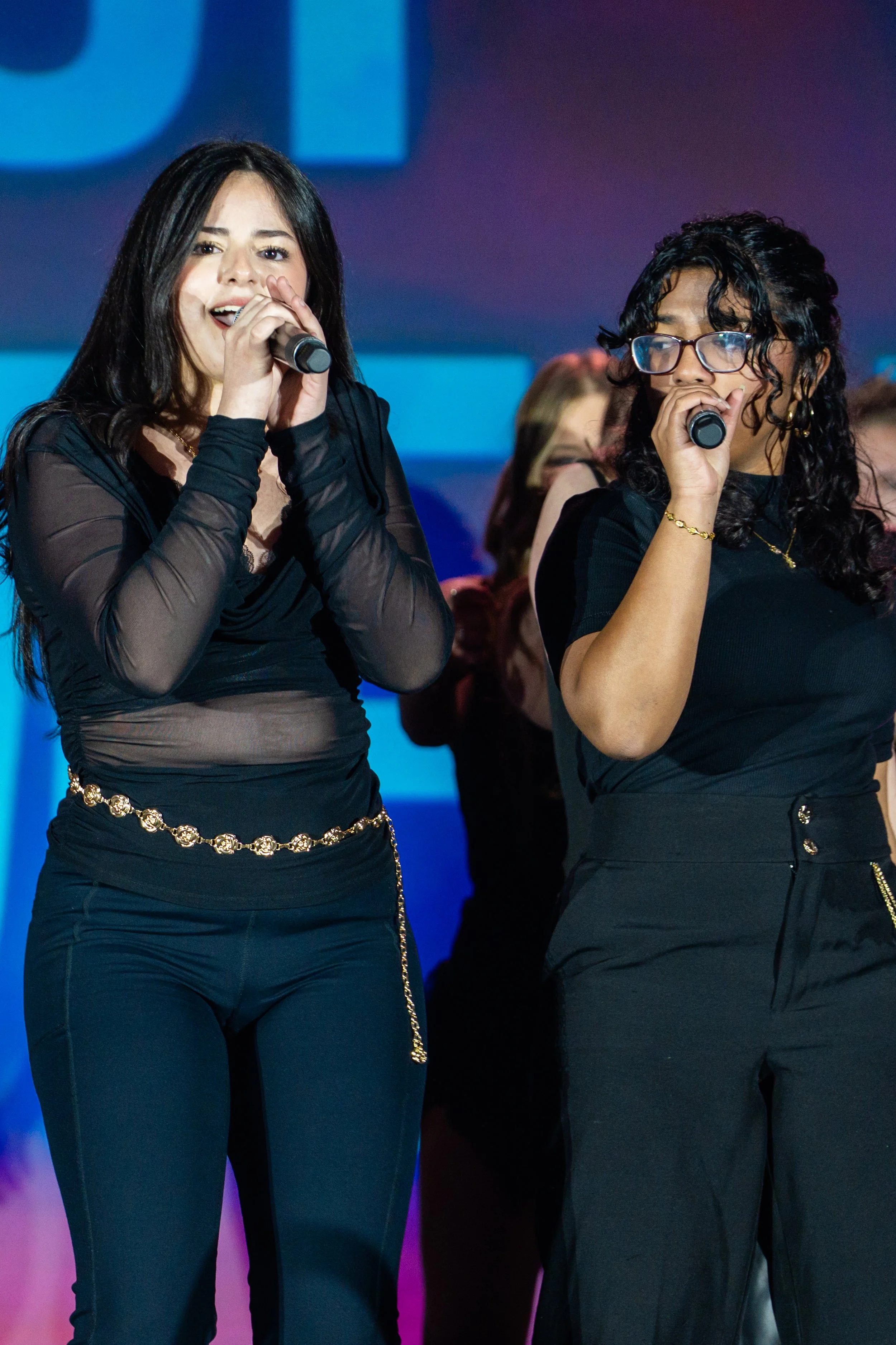 Two women singing with microphones on stage, one with long dark hair and sheer black top, the other with curly dark hair, glasses, and black dress, in front of a colorful background.