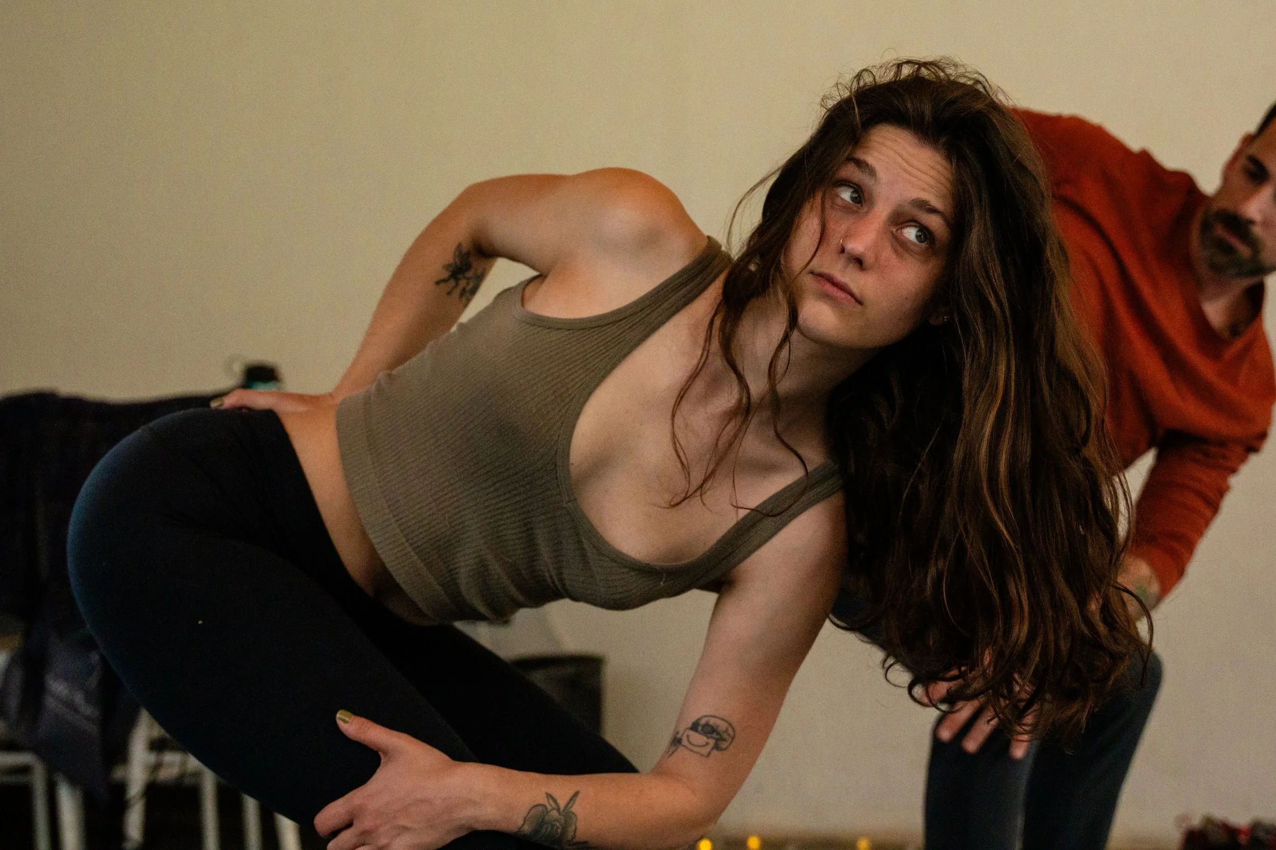 A young woman with long, curly brown hair, light skin, multiple tattoos, and a nose ring, practicing yoga or stretching in a room with beige walls, possibly with an instructor or fellow participant in the background.