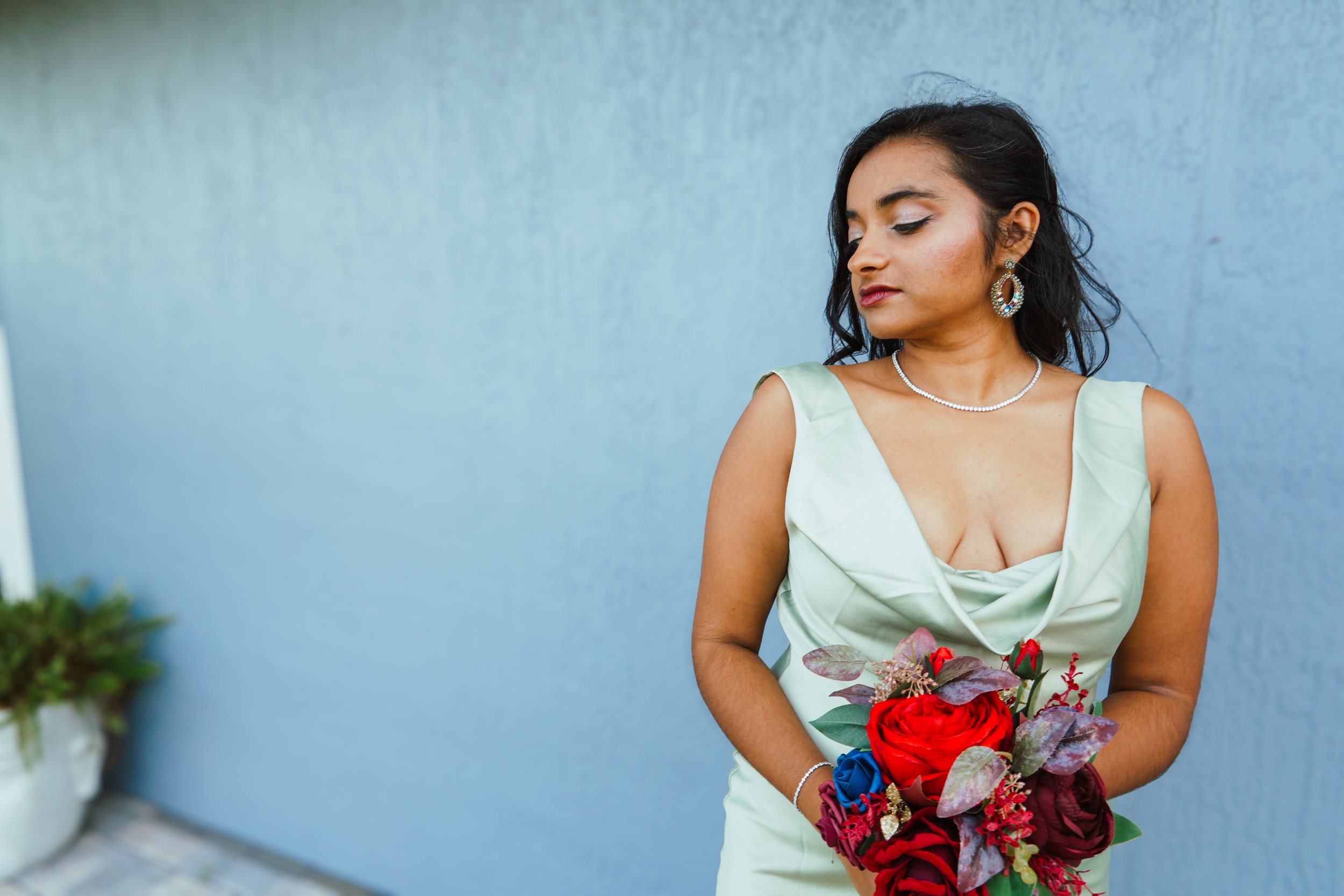 A woman with dark hair wearing earrings, a pearl necklace, and a light green dress holding a bouquet of red and purple flowers, standing against a gray wall.