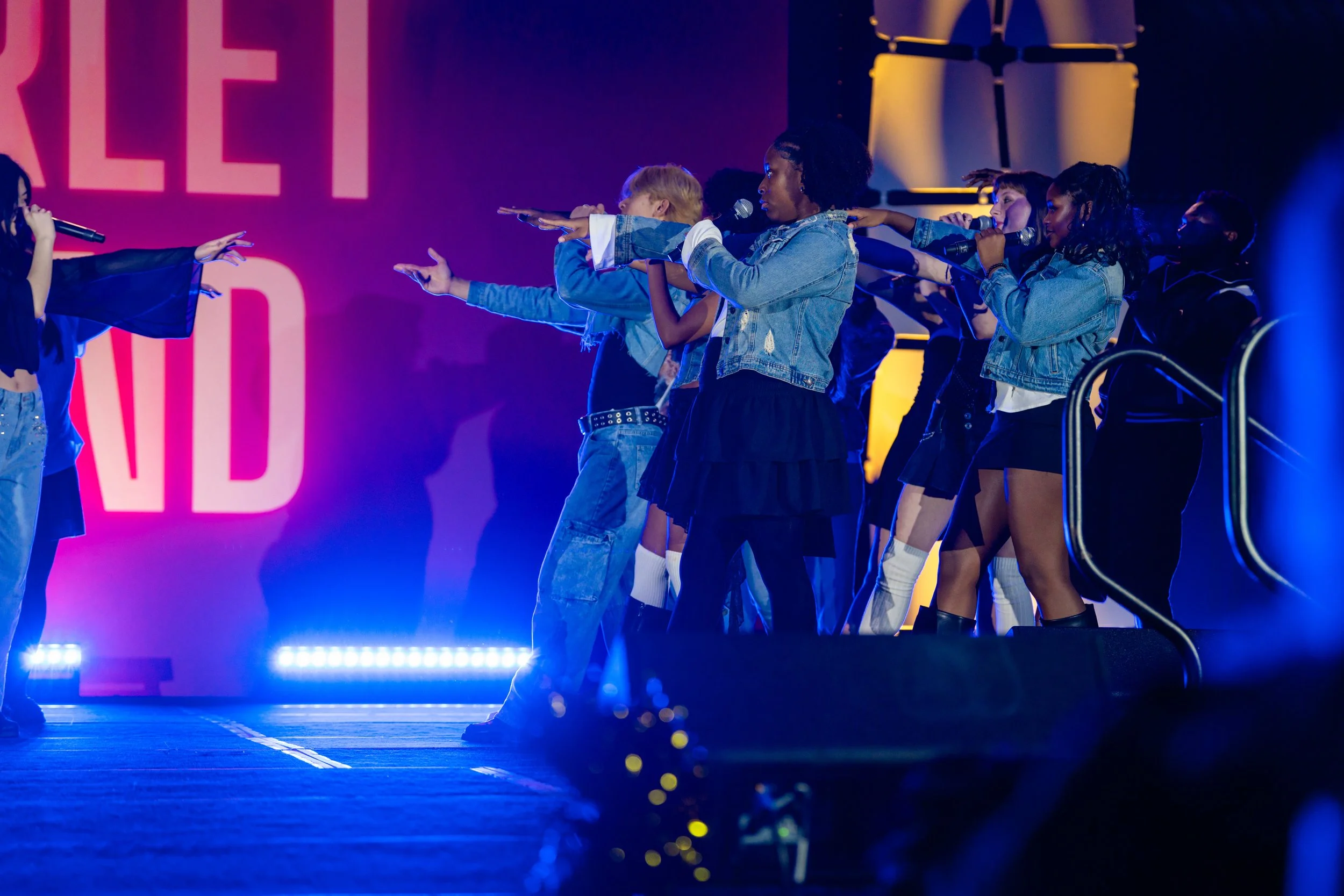 A group of women on stage, participating in a performance or rehearsal, pointing towards a woman who is speaking into a microphone, with colorful lights and a large pink sign with partial letters in the background.