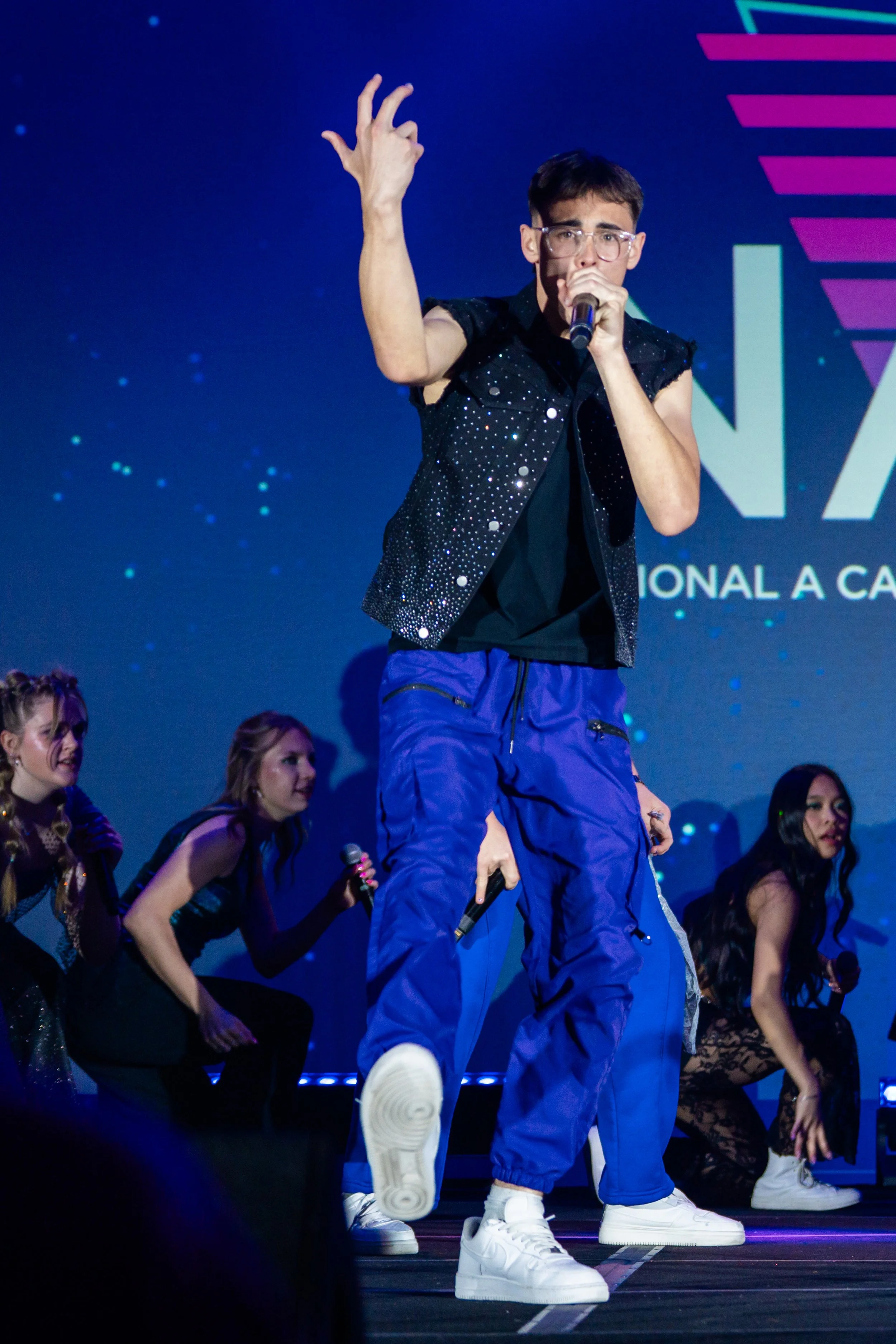A performer with short dark hair, glasses, and a black glittery shirt singing into a microphone on stage with backup dancers in sporty outfits.