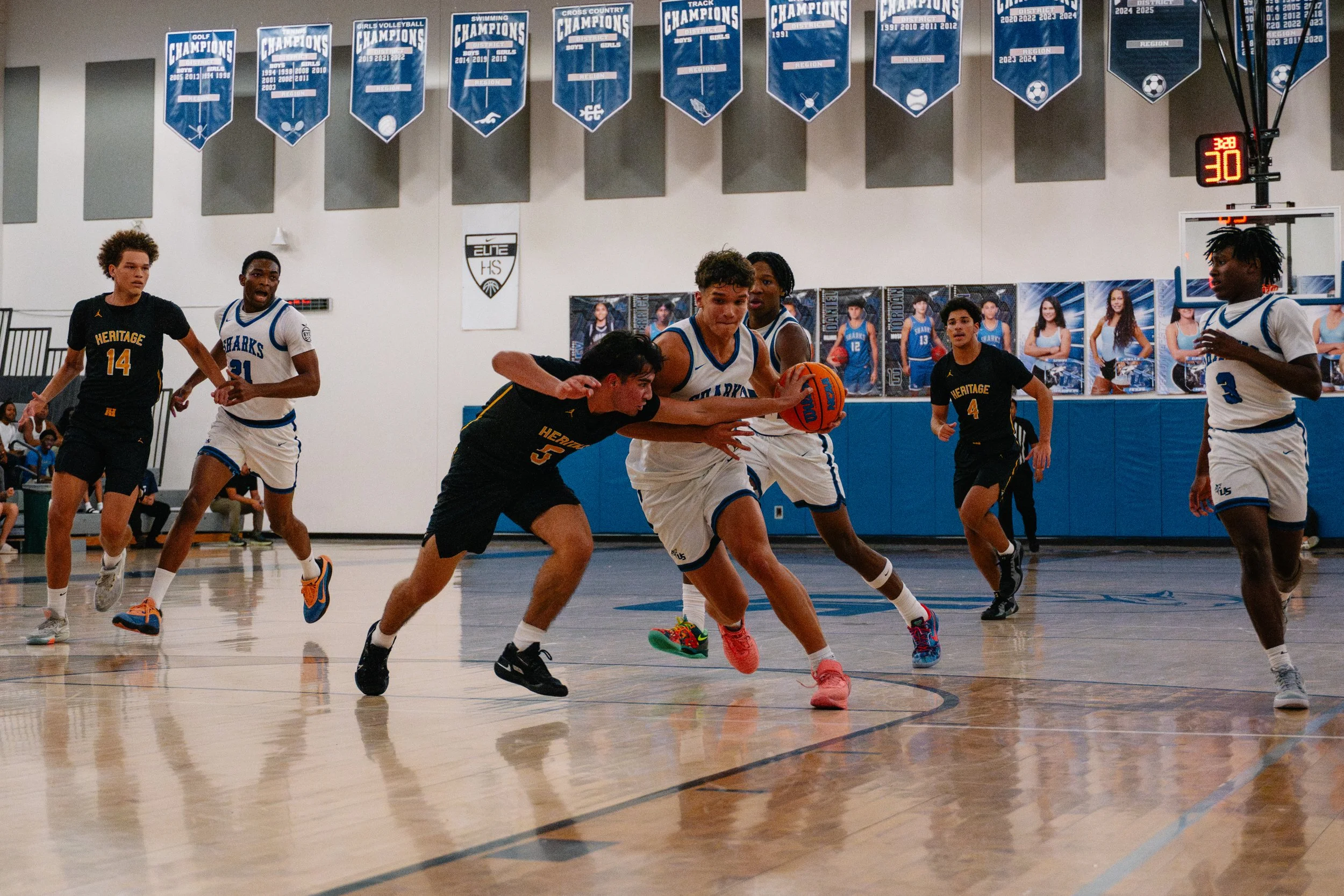 A competitive basketball game between two teams, one in black and yellow uniforms and the other in white and blue uniforms, with players trying to gain control of the ball on the court.
