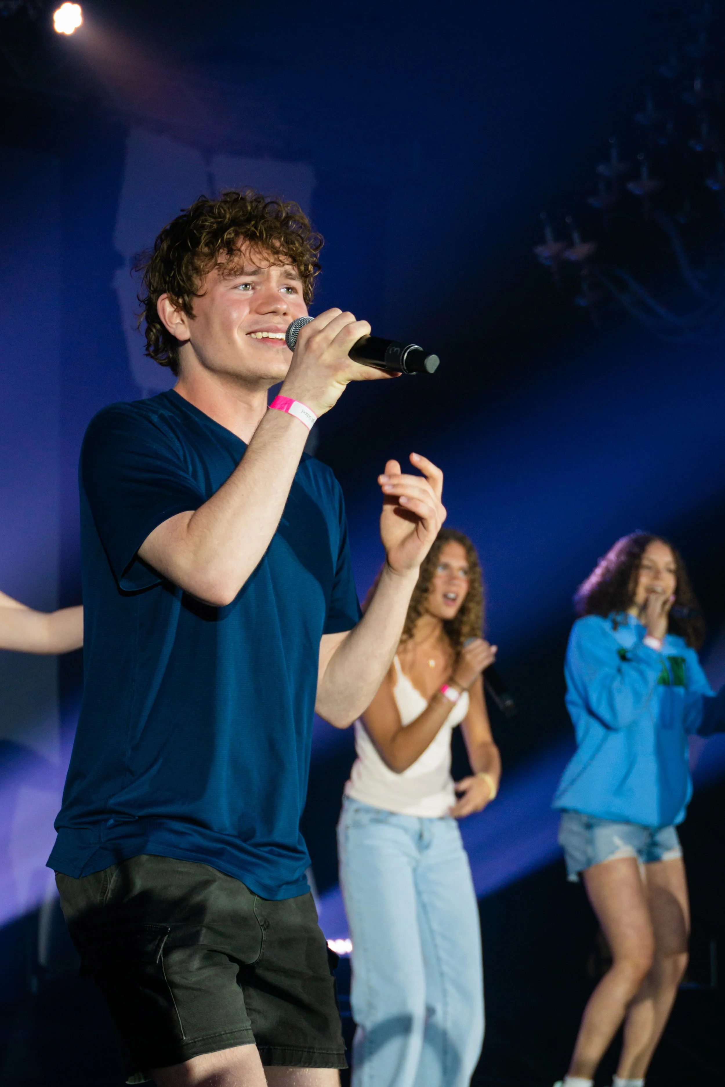 A young man with curly hair and a pink wristband holding a microphone on stage, smiling, with three women in casual outfits singing and dancing in the background under stage lights.