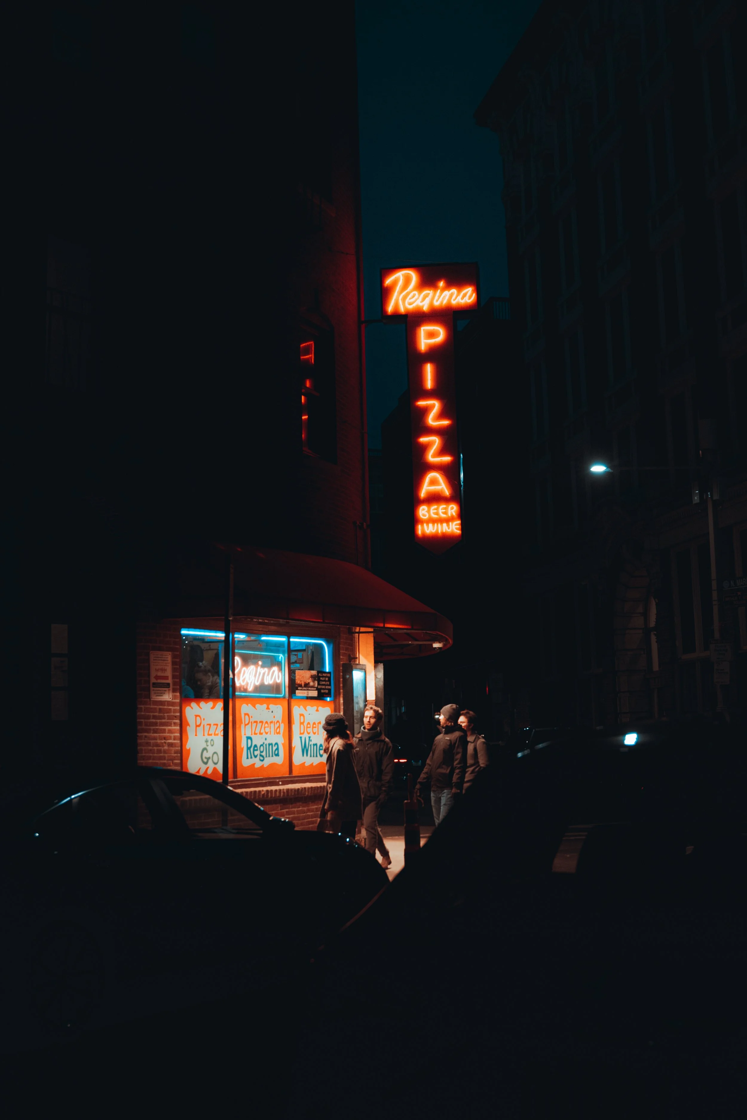 Nighttime street scene featuring a pizzeria with neon signs that read "Regina" and "Pizza to Go." Several people stand outside the pizzeria, and a few cars are parked nearby.