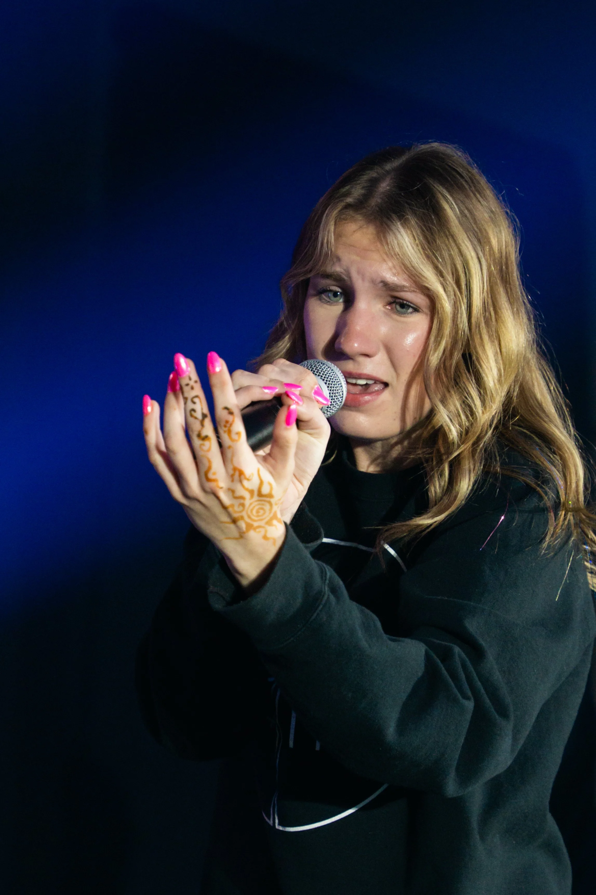 A young woman with wavy blonde hair and light skin singing into a microphone. She has pink painted nails, henna-style hand art, and is wearing a black hoodie. The background is dark blue.