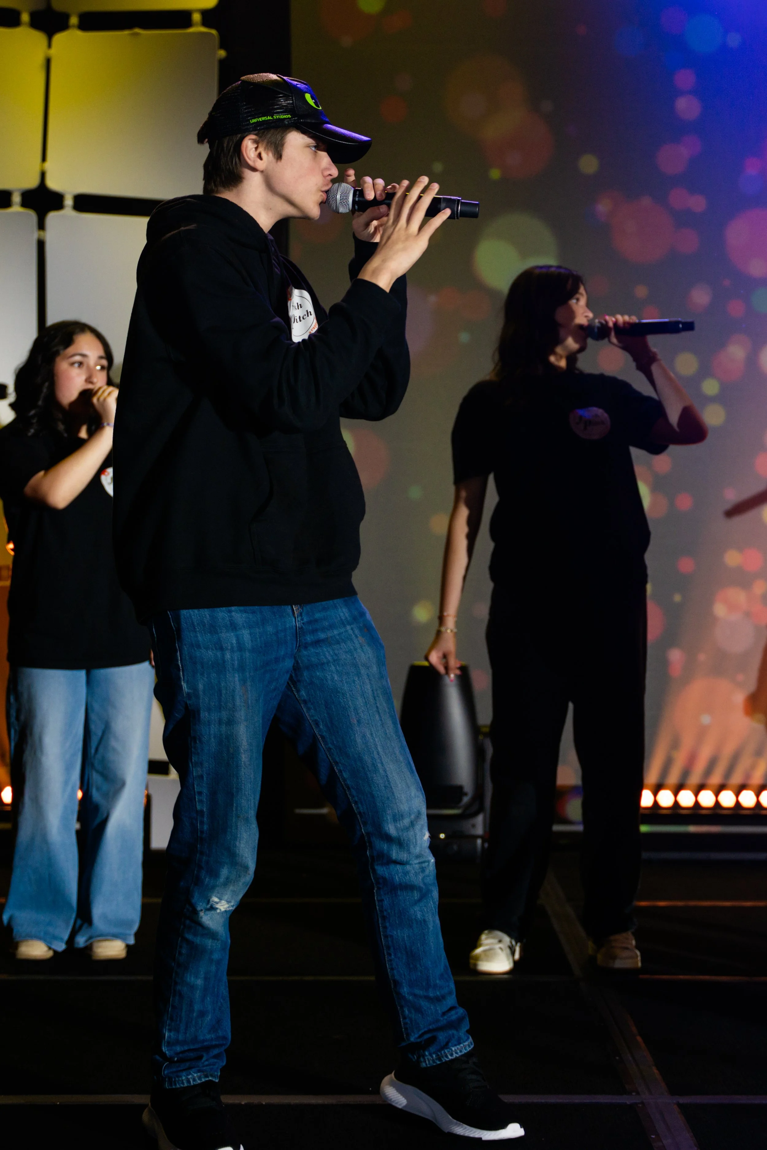 A group of young people performing on stage, with a young man in the foreground singing into a microphone and wearing a black hoodie and blue jeans, while others in the background sing into microphones.