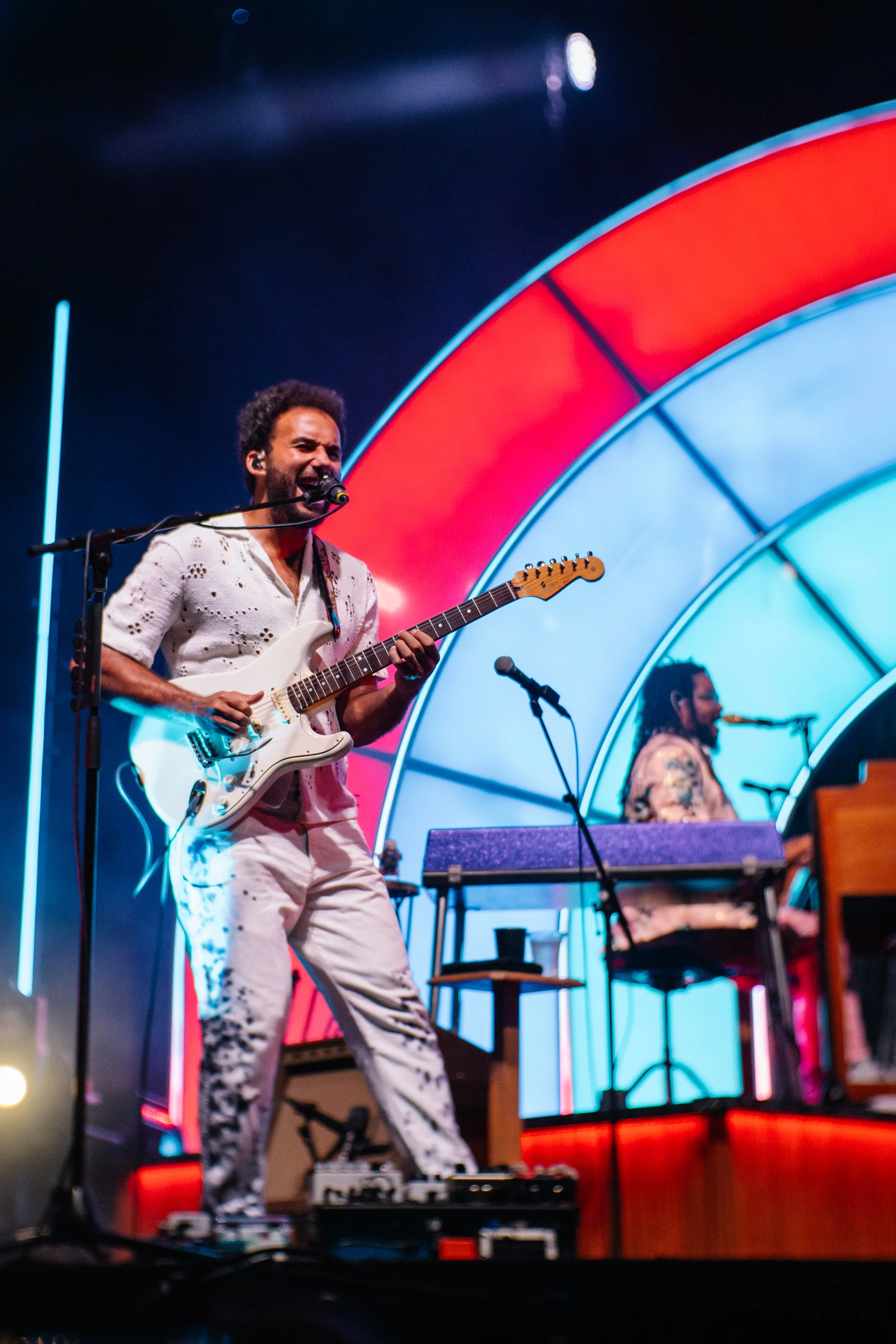 Musician playing electric guitar and singing into microphone on stage with colorful light display in background.