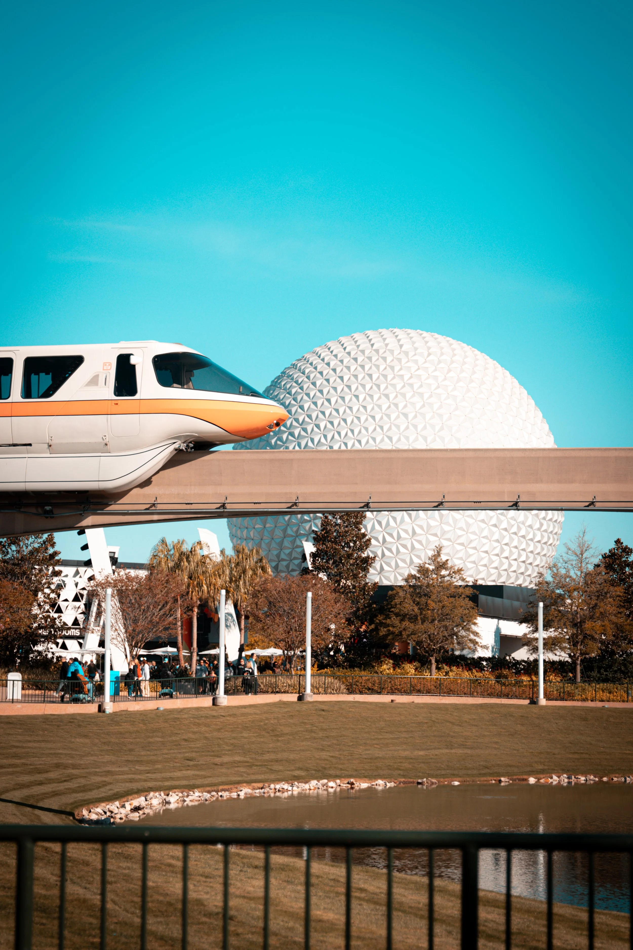 A monorail train passes in front of Spaceship Earth at Epcot theme park in Disney World with clear blue sky above and trees lining the park.