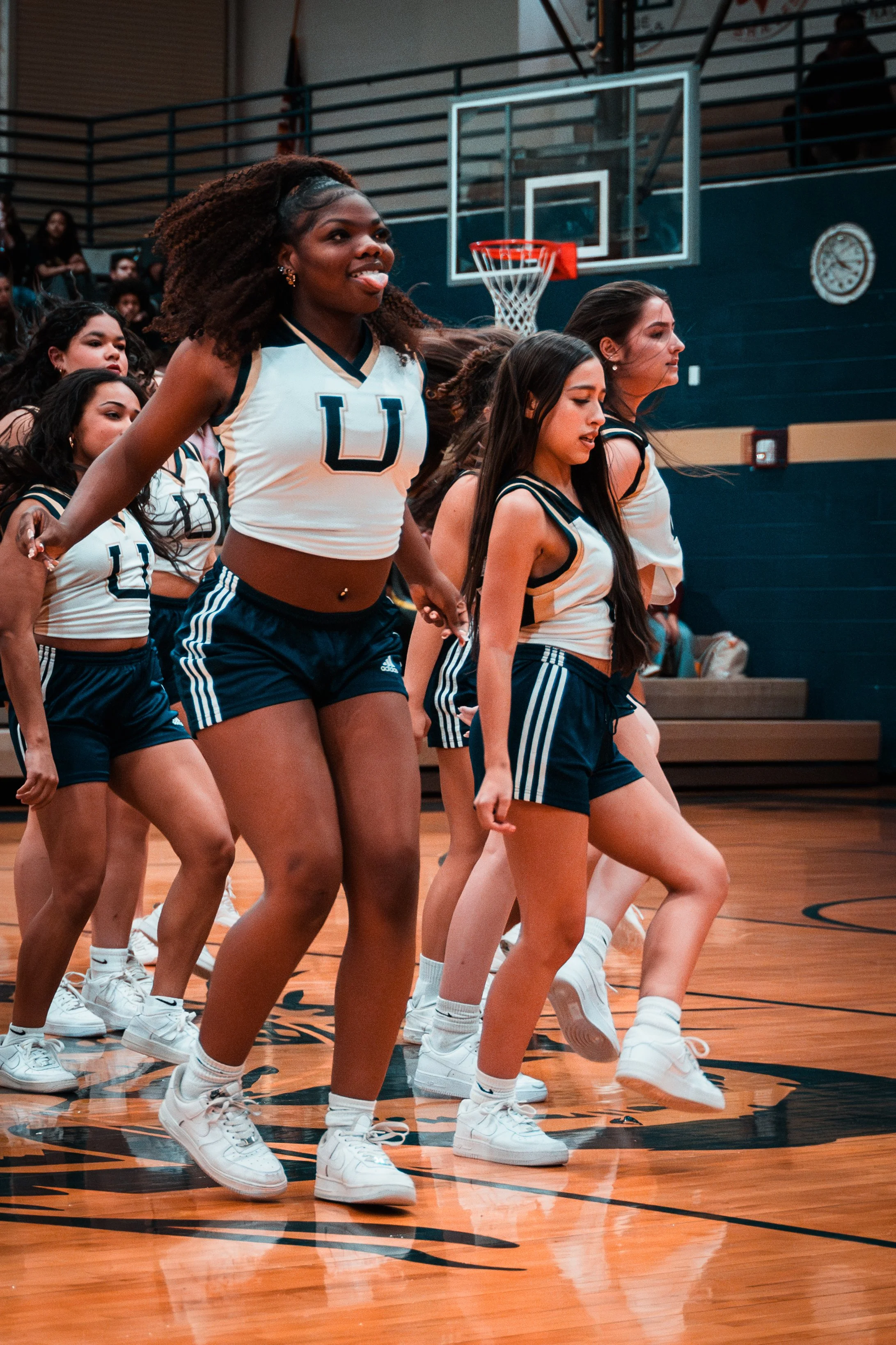 A group of young women cheerleaders in matching white and navy sports uniforms, performing a routine on a basketball court with a basketball hoop and spectators in the background.