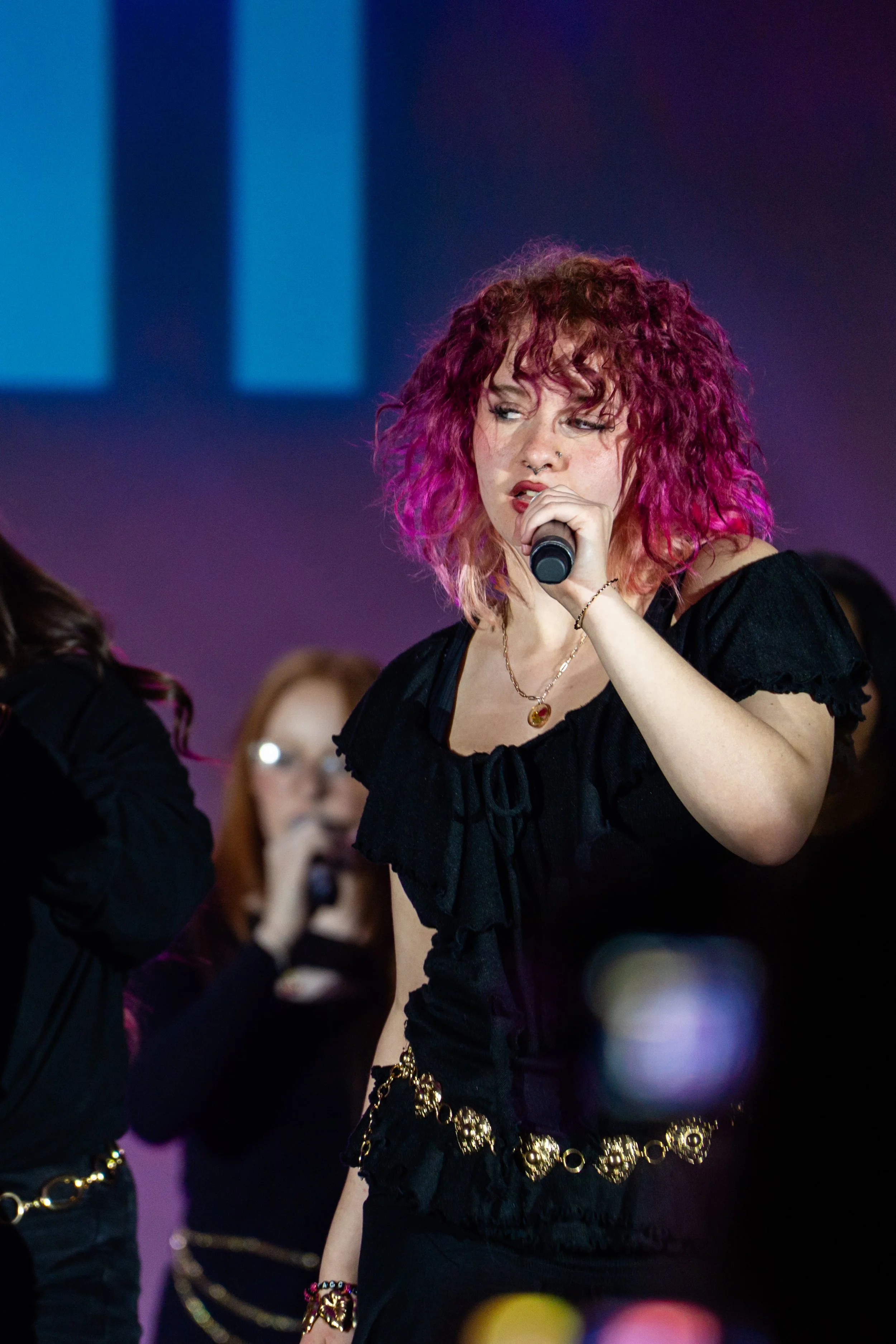 Young woman with curly pink and purple hair singing into a microphone, wearing a black top and gold jewelry, on stage with a blurred background.