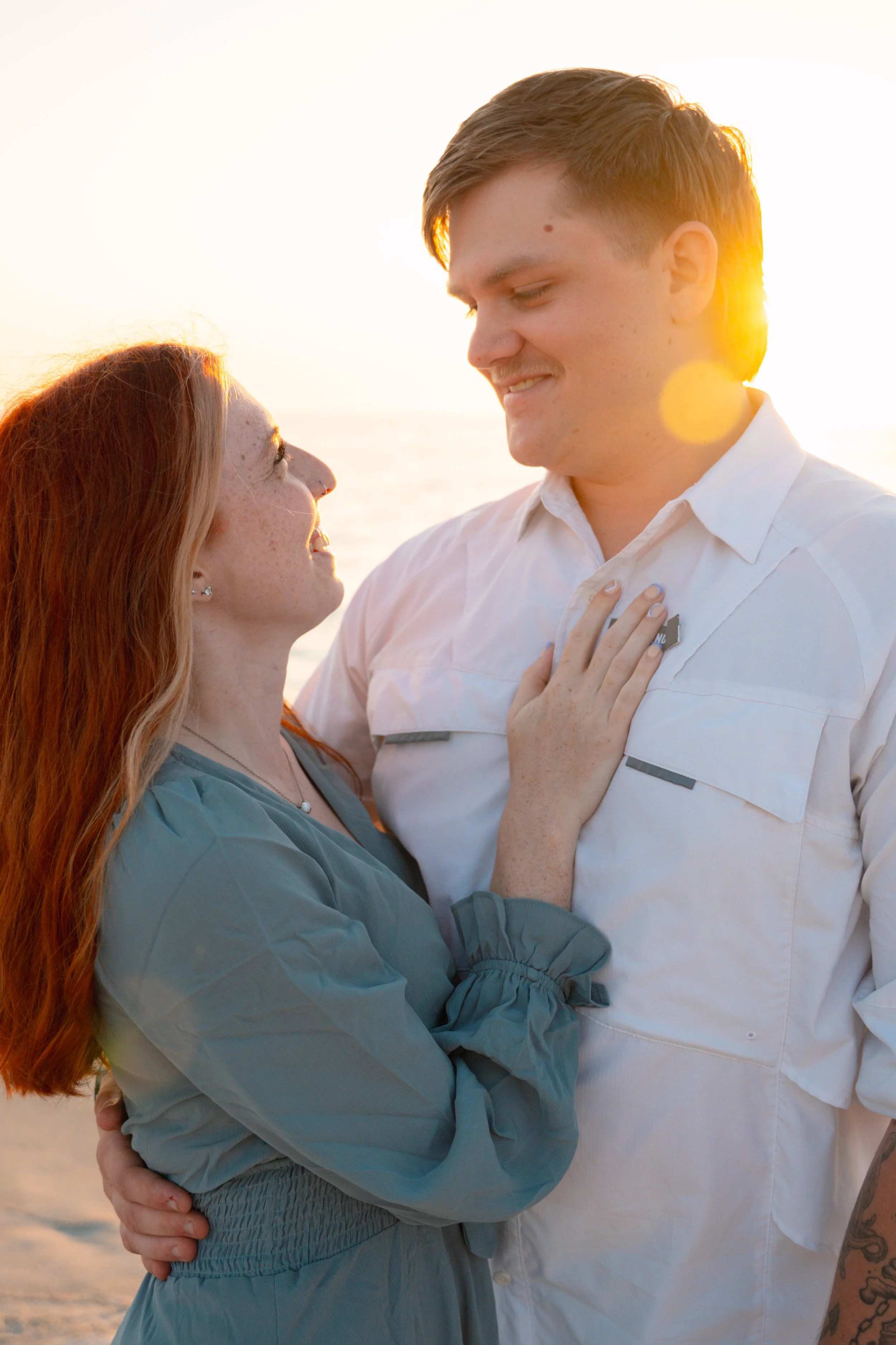 A couple standing close together on a beach at sunset, gazing smiling into each other's eyes.