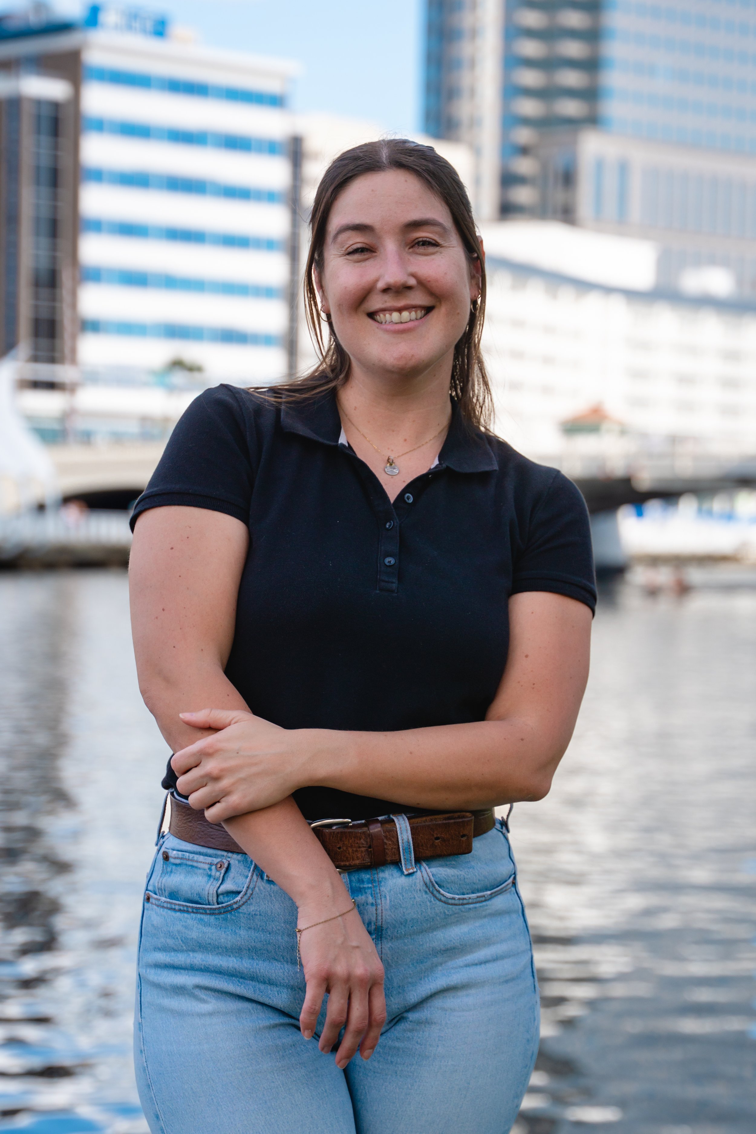 A woman smiling with arms crossed, standing near a body of water with modern office buildings in the background.