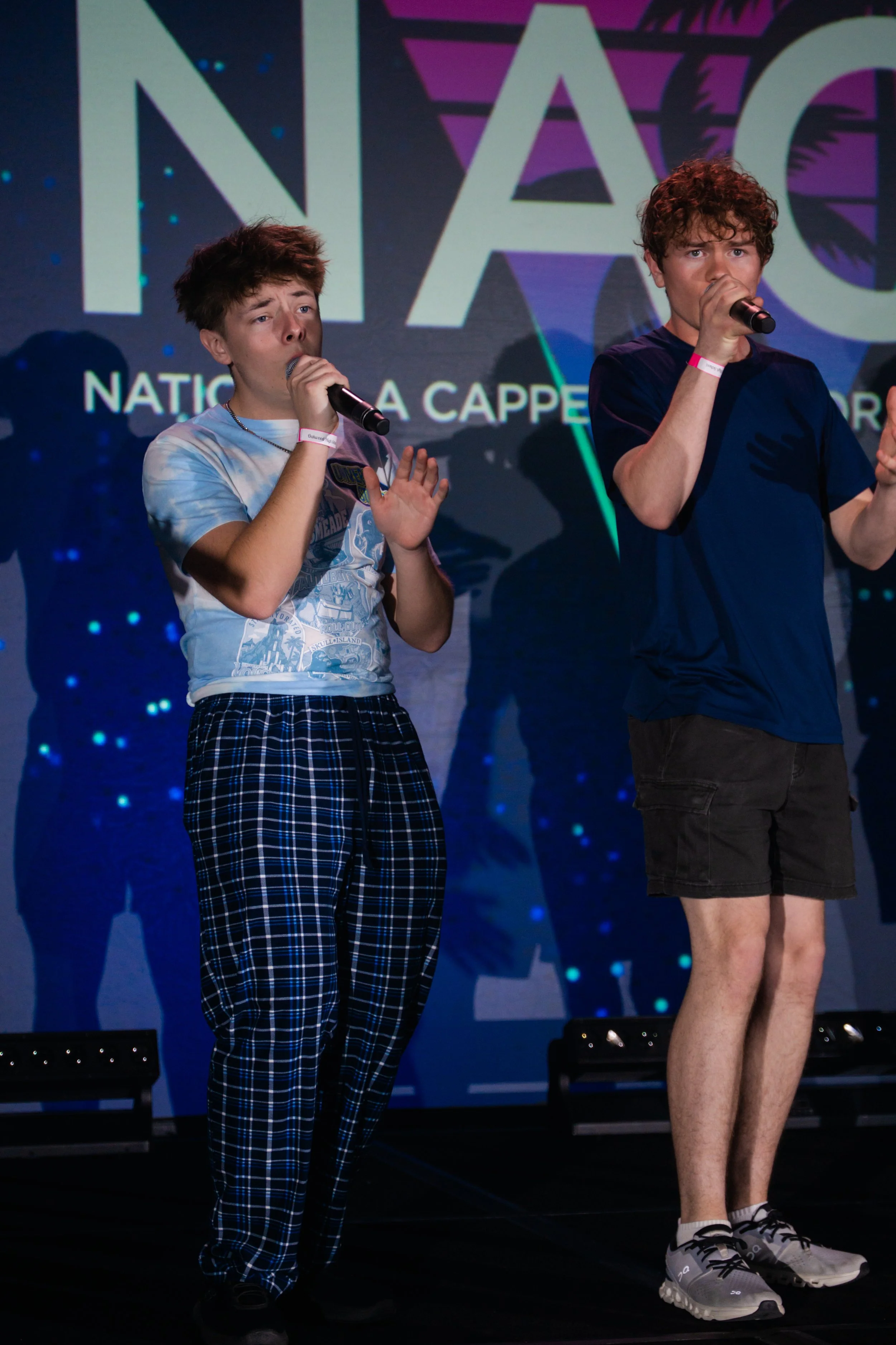 Two young men standing on a stage, singing into microphones, with a backdrop displaying the text 'NAC' and 'NATIONAL' in large letters.