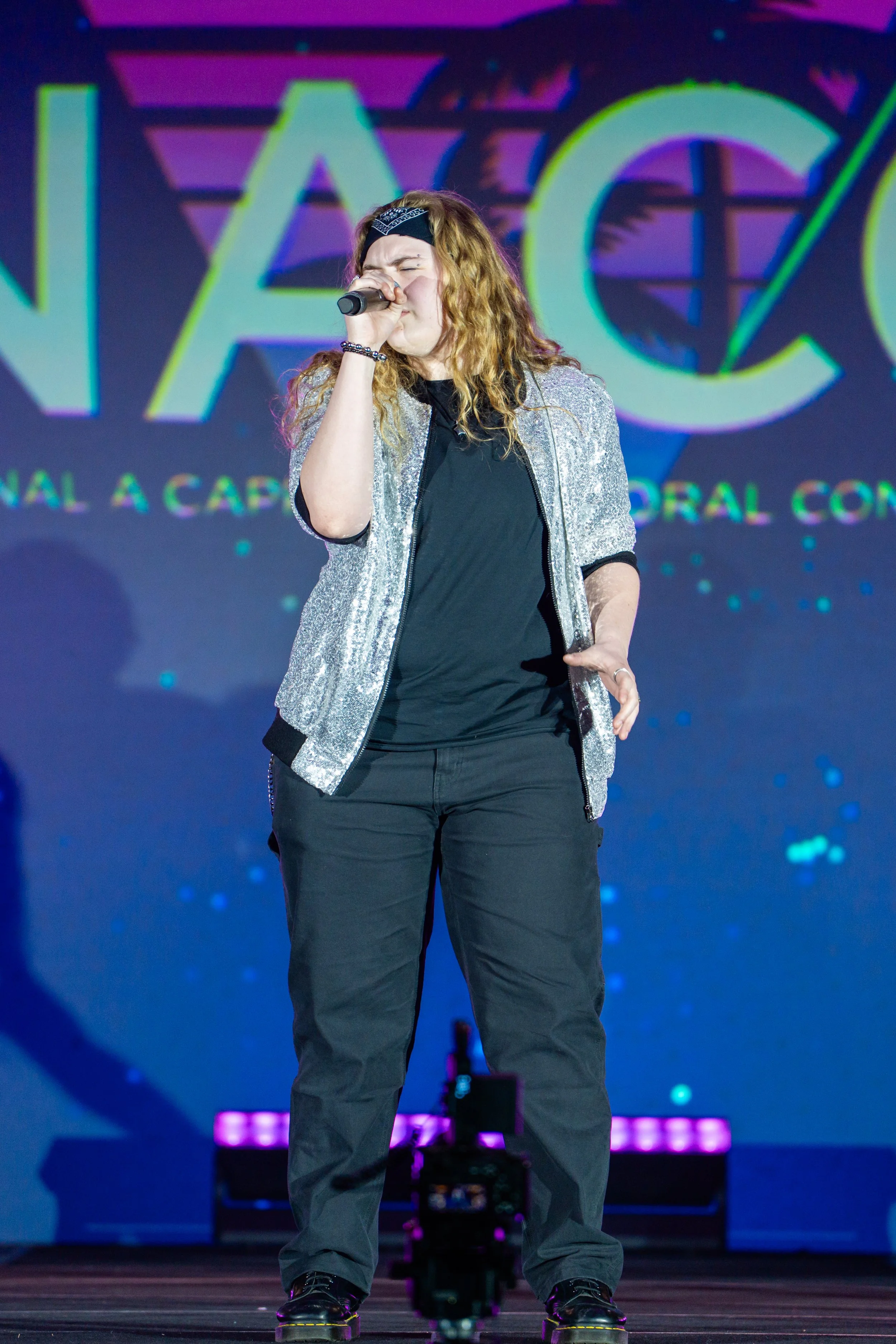 A woman with curly hair, wearing a bandana, black shirt, gray jacket, and black pants, singing into a microphone on stage with a colorful background.